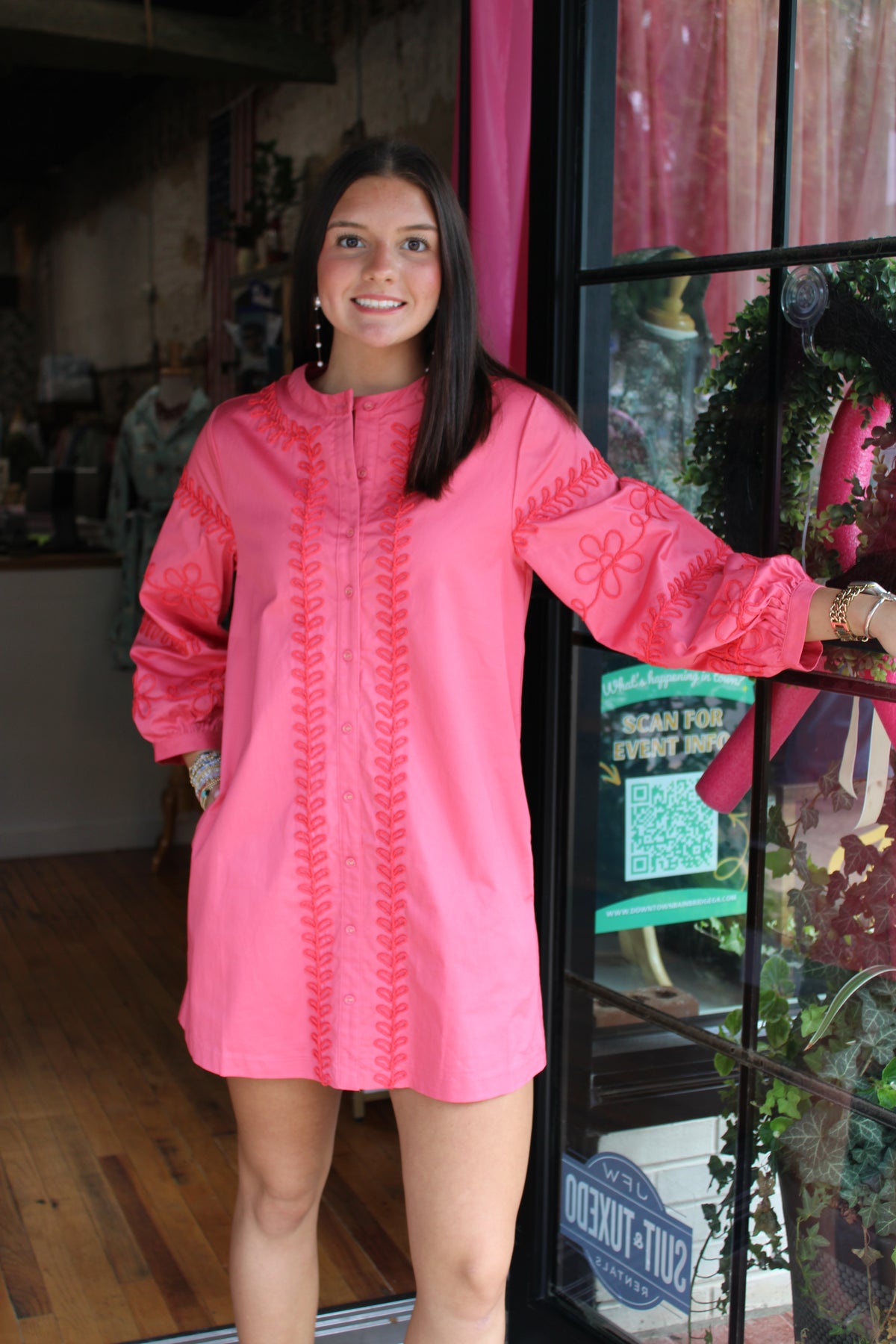 A woman wearing a bright pink long-sleeve tunic dress with tonal pink embroidery, standing in front of a shop window.