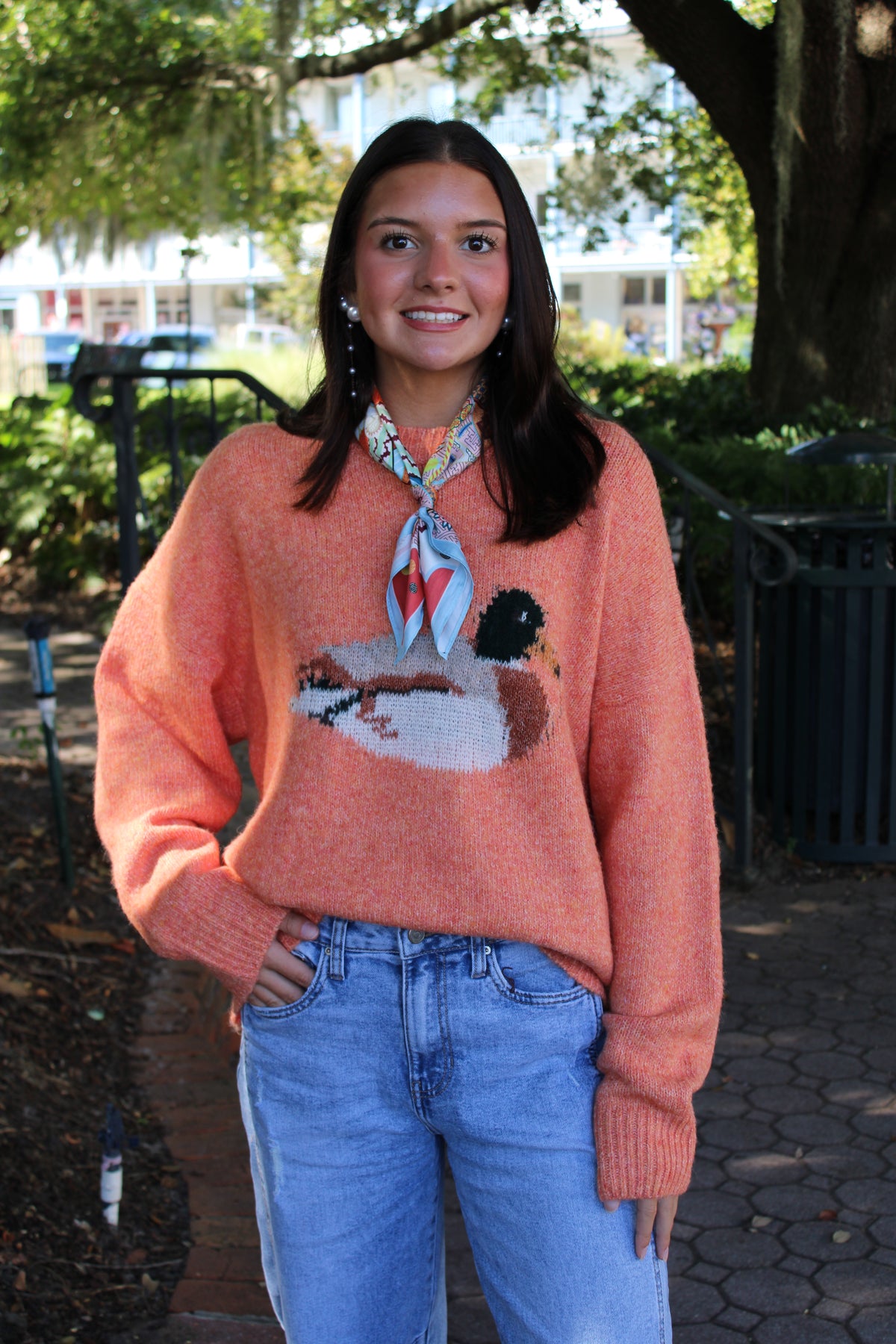 A woman wearing a fuzzy coral-orange crewneck sweater with a large duck graphic on the front, styled with a patterned neck scarf and light-wash blue jeans, standing outdoors on a paved path with trees in the background.