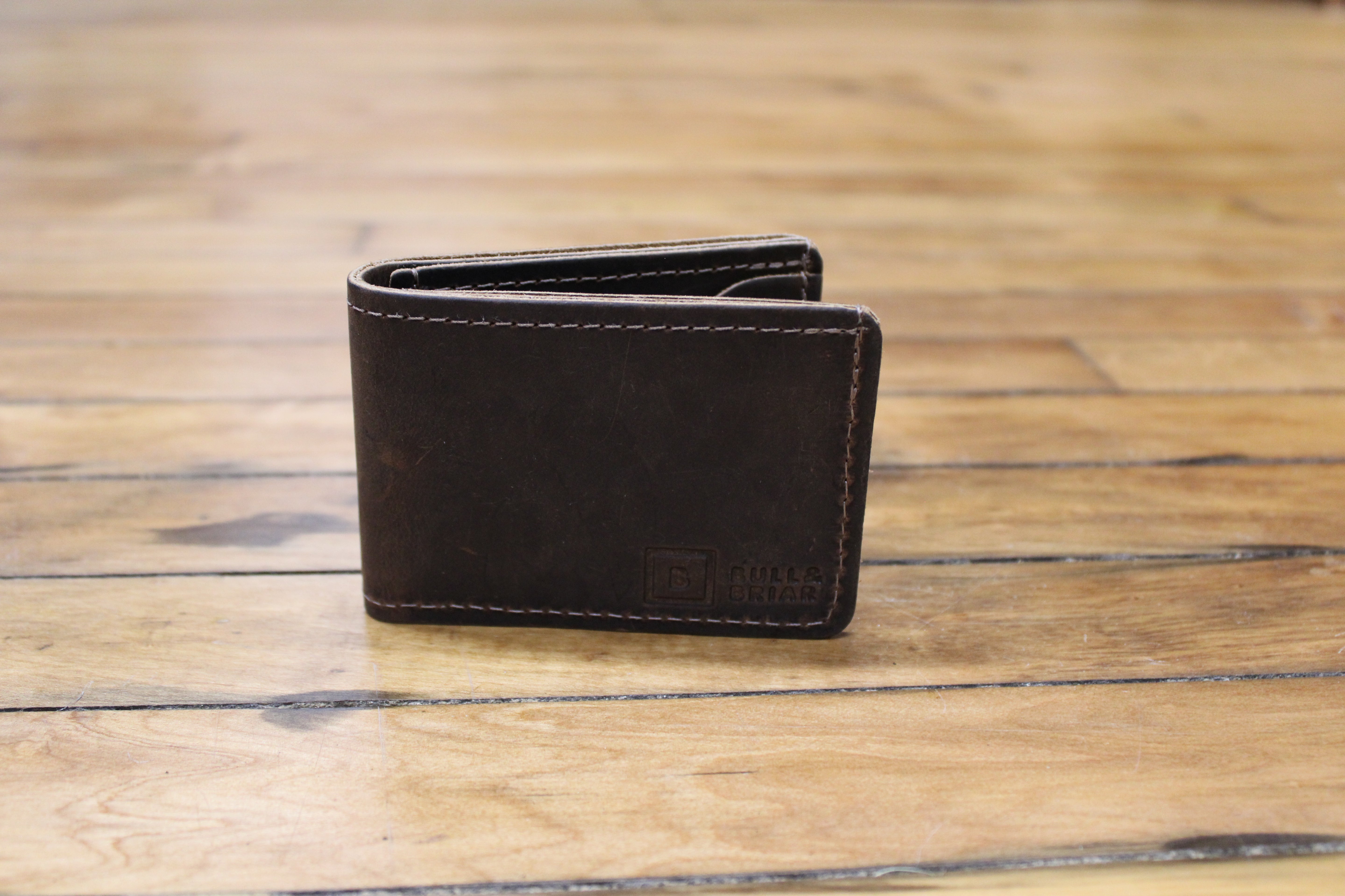 A dark brown leather bifold wallet with visible stitching and a debossed logo on the front