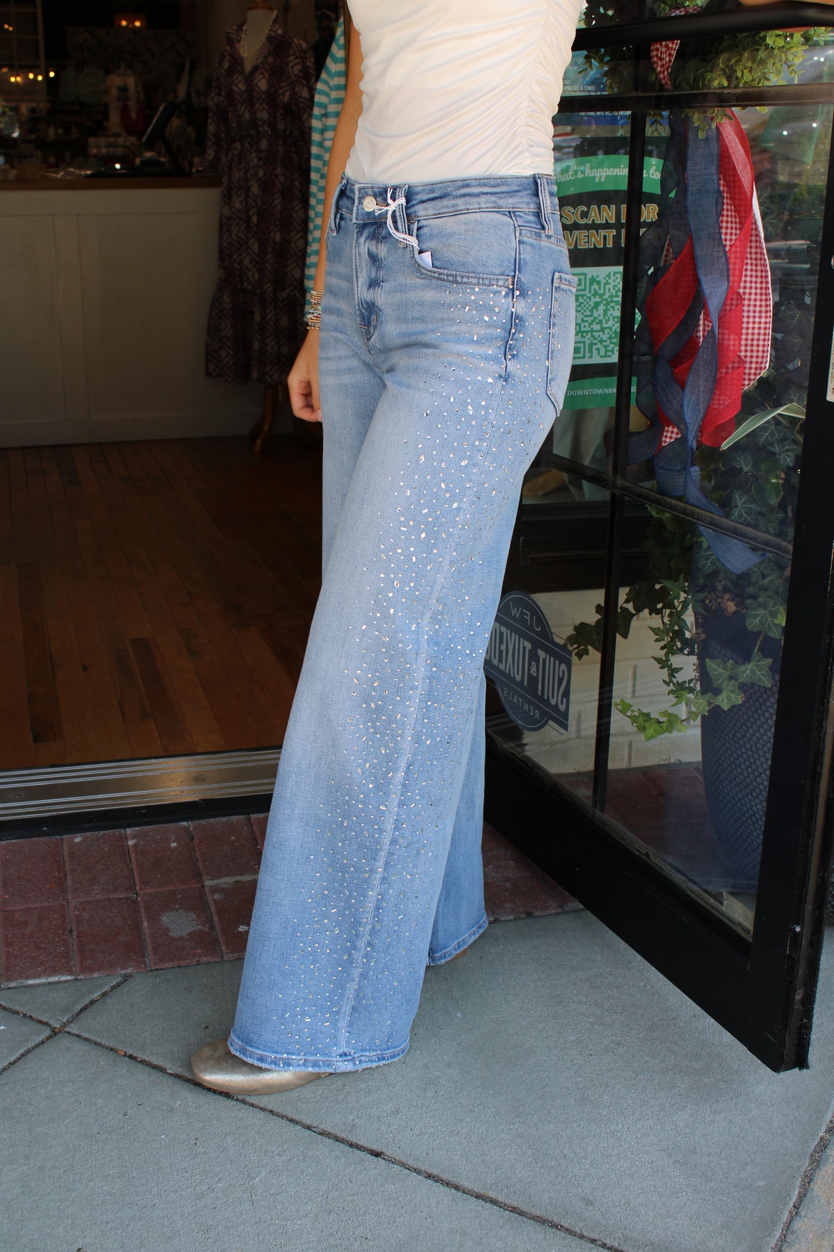 A person wearing light-wash blue wide-leg jeans with small silver rhinestone embellishments, standing on a sidewalk in front of a storefront.