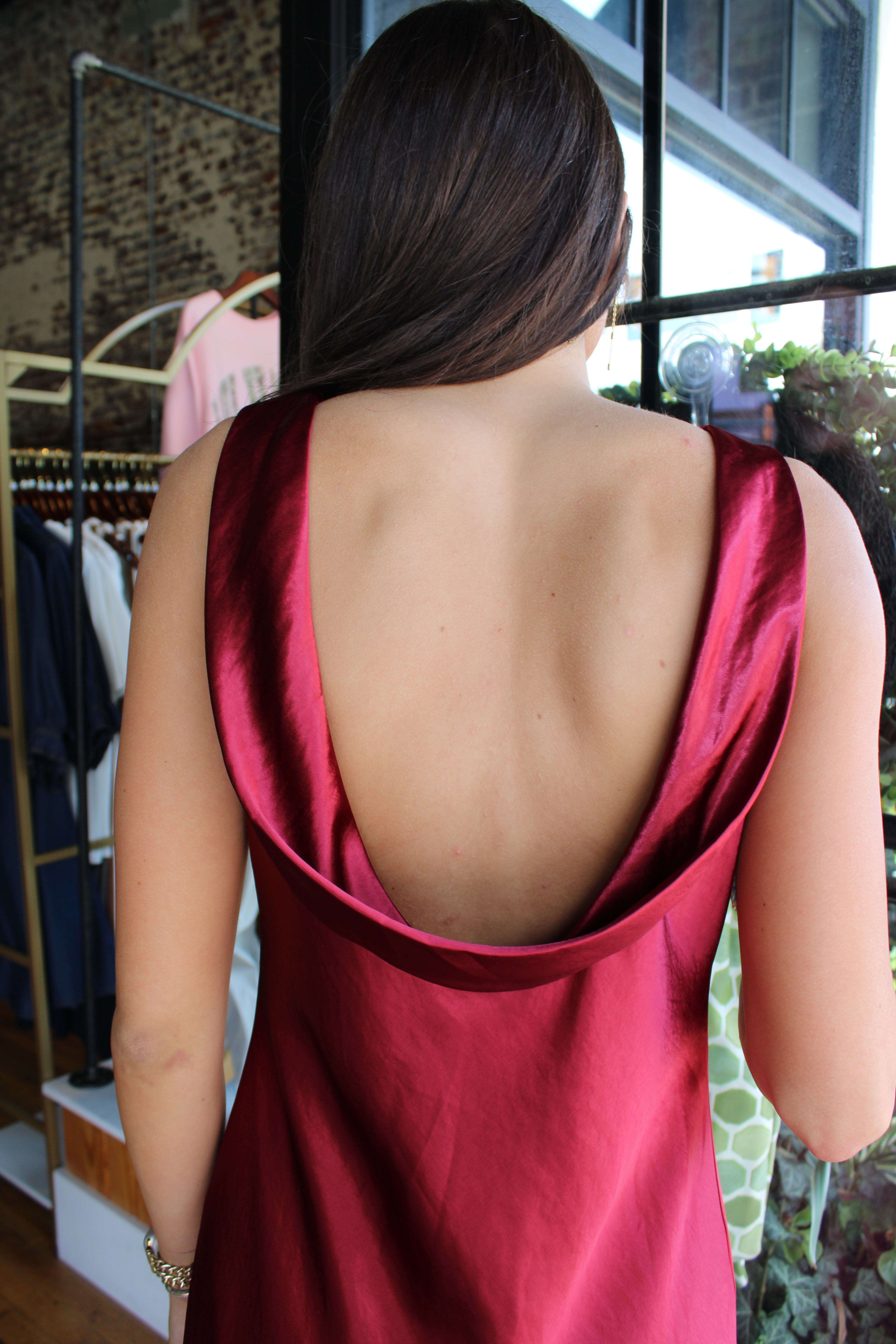 A woman viewed from behind wearing a sleeveless satin slip dress with a very large open back in a deep wine red color, set in a boutique environment.