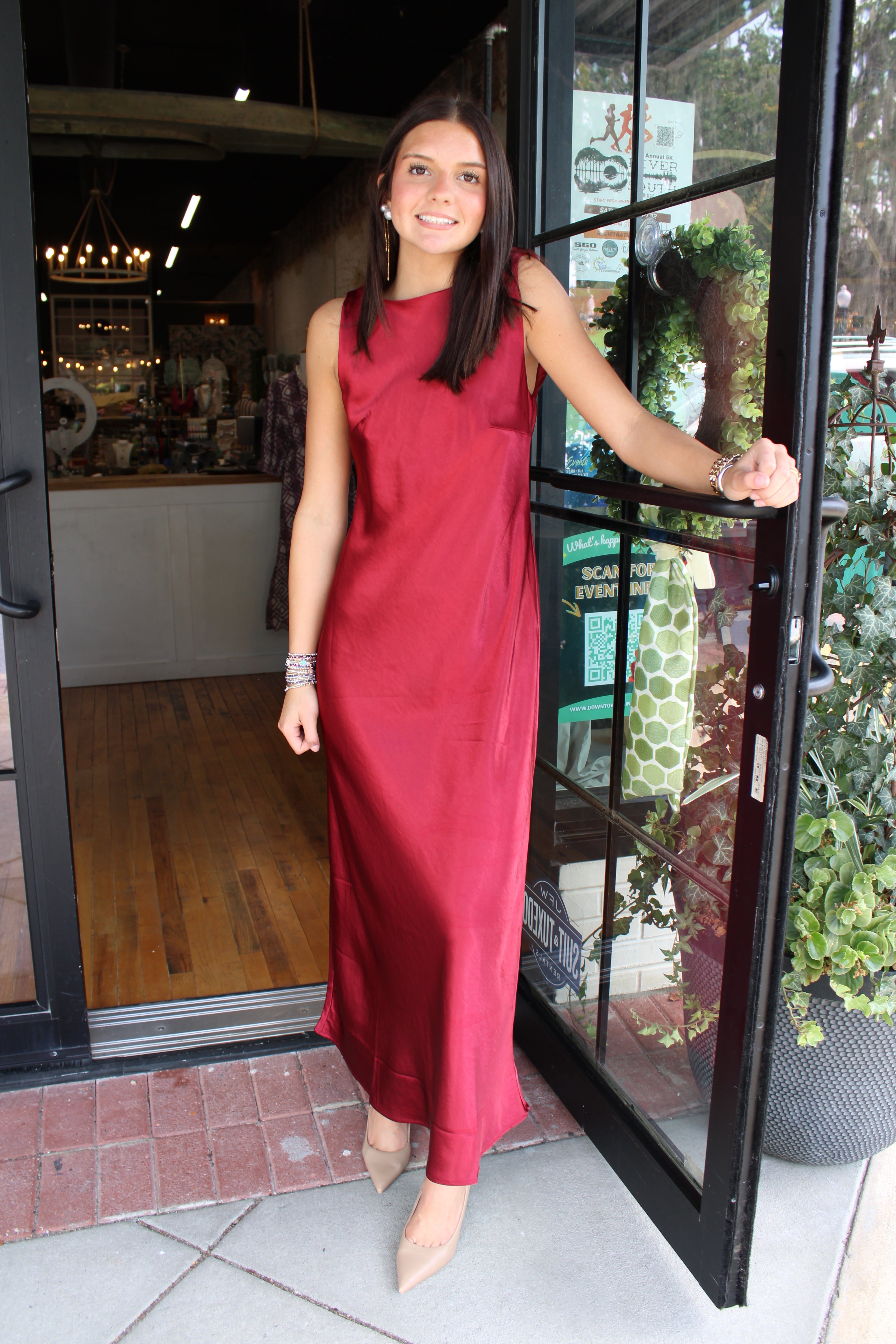 A woman wearing a sleeveless, high-neck, floor-length red dress and beige pointed-toe heels stands in an open storefront doorway