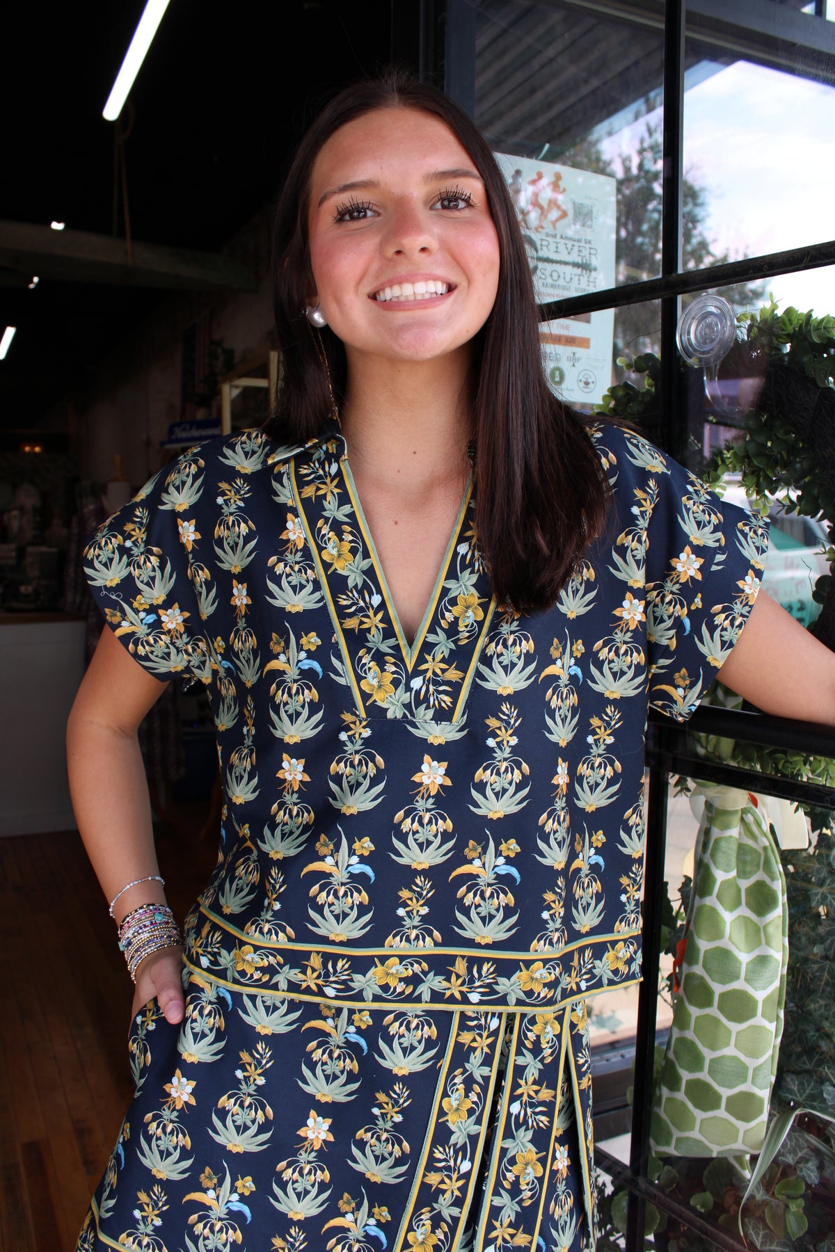 A smiling young woman with long dark hair stands indoors wearing a navy blue short-sleeve set with a yellow and light blue floral pattern and a matching belt. She is posed in front of a window in what appears to be a boutique setting.