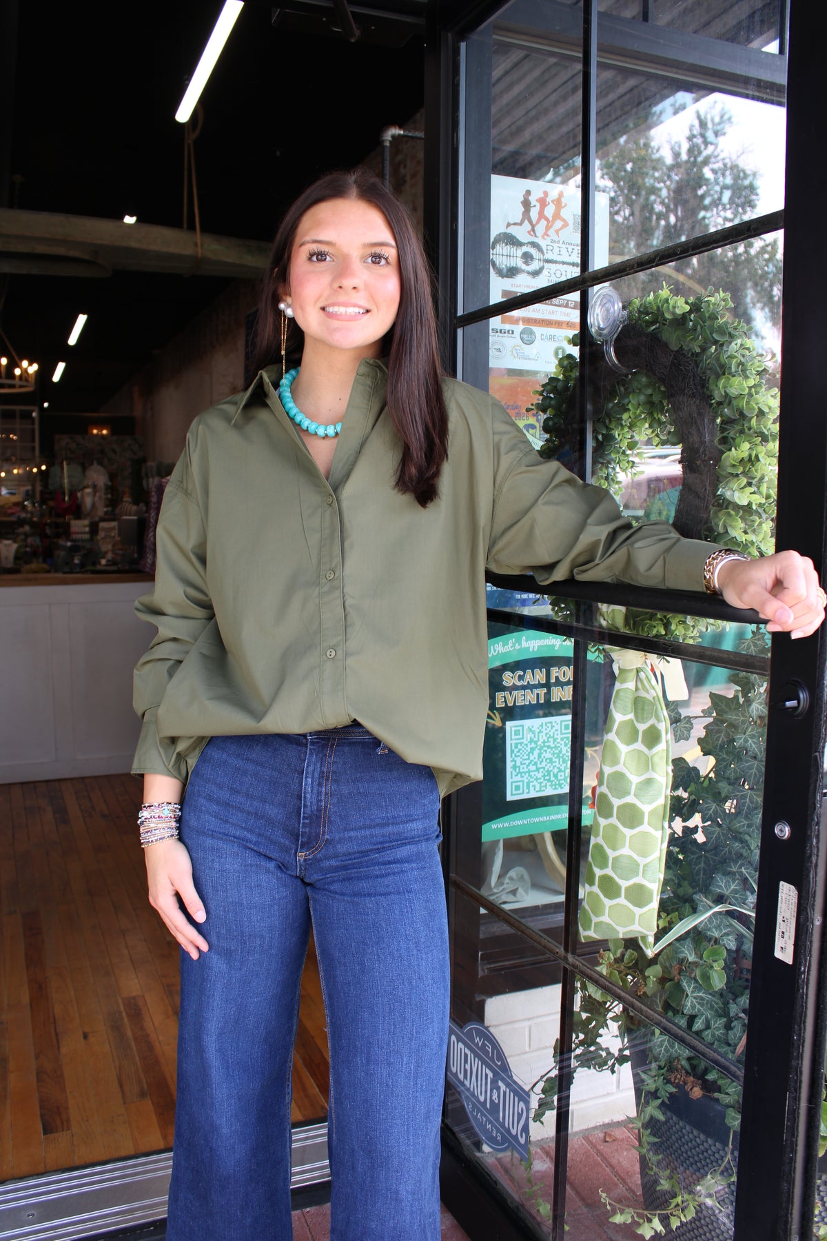 A woman stands in a storefront doorway wearing an olive green long-sleeve button-up shirt and blue jeans, with a turquoise necklace and bracelets visible.