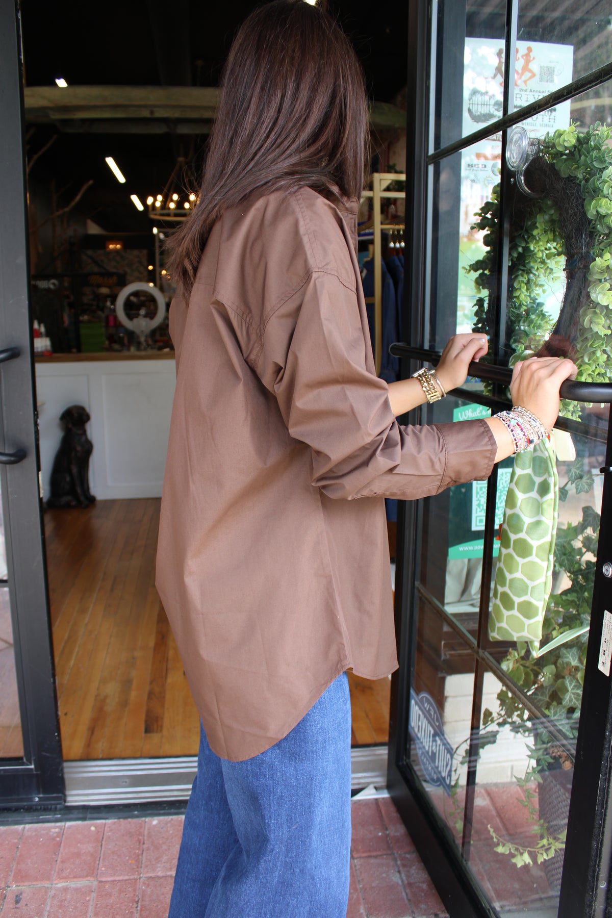 A woman wearing an oversized brown long-sleeve shirt and blue jeans stands at a glass storefront door, holding the handle. The photo is taken from behind, showing the back of the shirt and her hair.