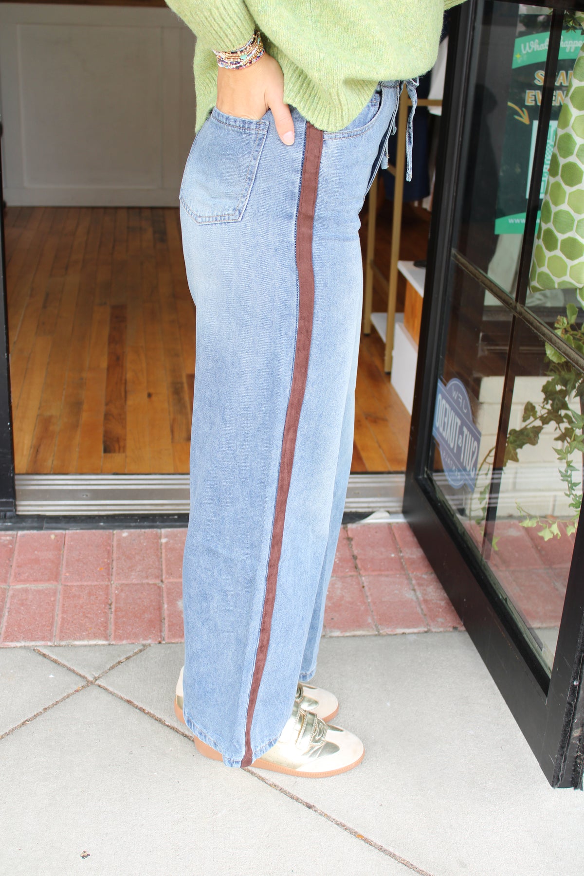 A person wearing a light blue wide-leg denim skirt with a vertical brown stripe down the side, paired with a light green sweater and white sneakers, standing on a sidewalk in front of a storefront.