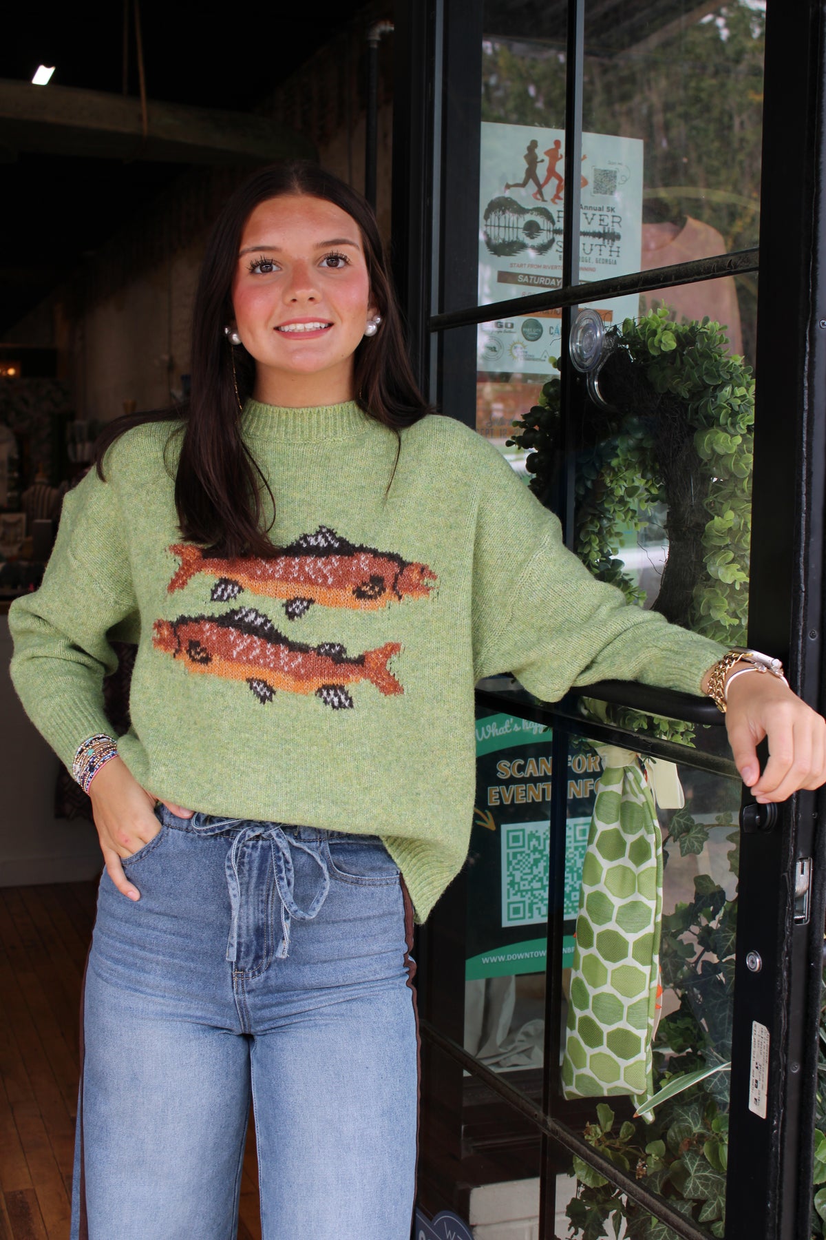 A woman wearing a light green knit sweater with two orange and black fish motifs on the front, paired with blue jeans, standing in front of a shop window.