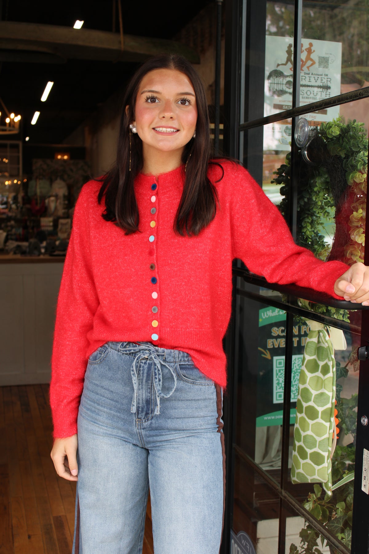 A woman wearing a bright red knit button-up sweater and light blue jeans stands in front of a storefront window, smiling at the camera.