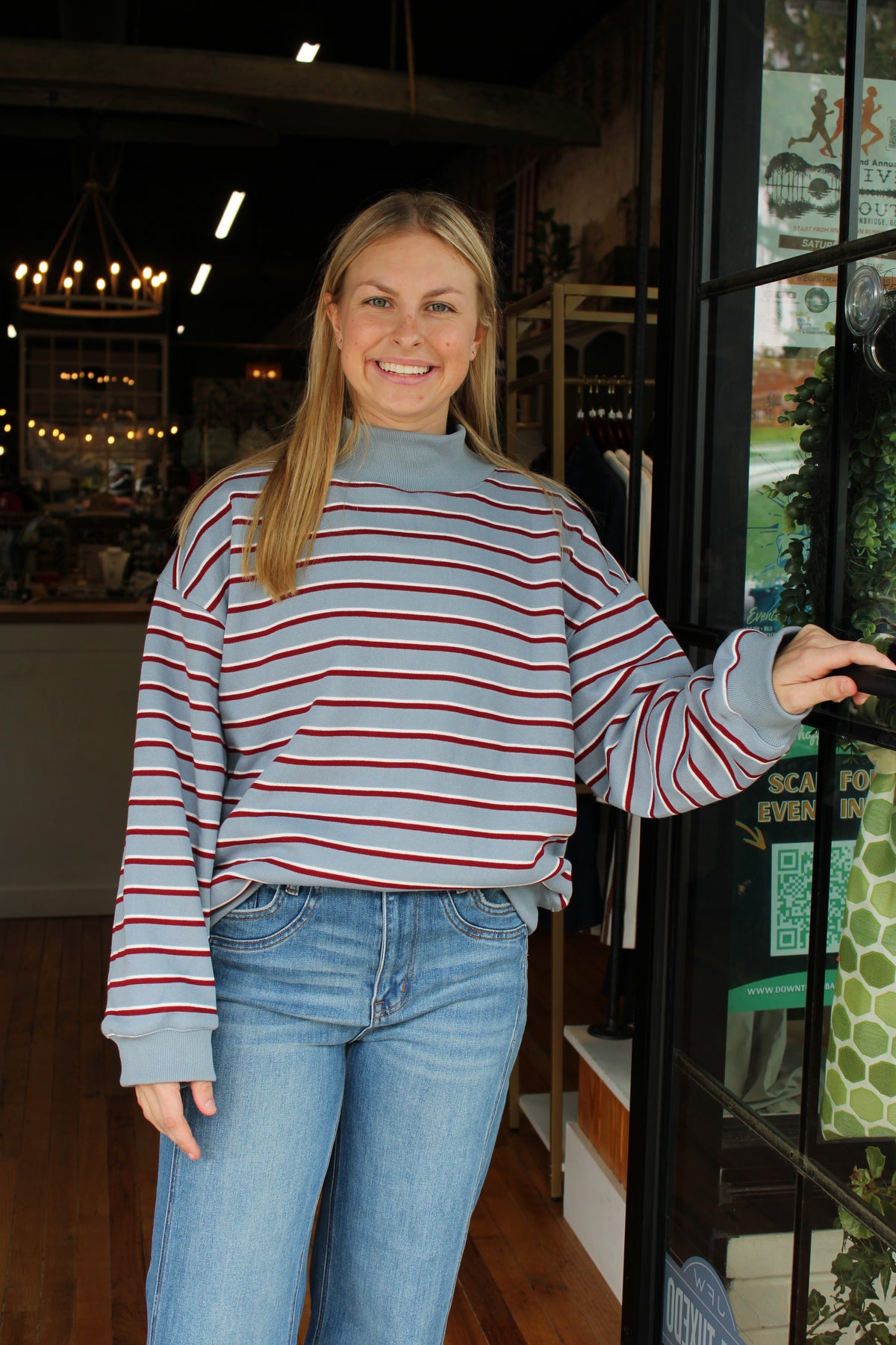 A woman with blonde hair stands in a shop doorway wearing a light blue mock-neck long-sleeve sweatshirt with thin red and navy horizontal stripes, paired with blue jeans.