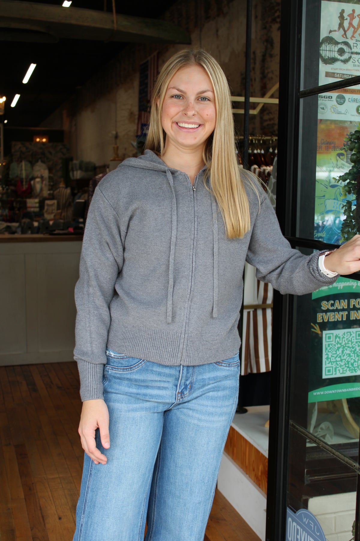 A smiling blonde woman wearing a gray zip-up hoodie and blue jeans stands in a retail store next to a glass door.
