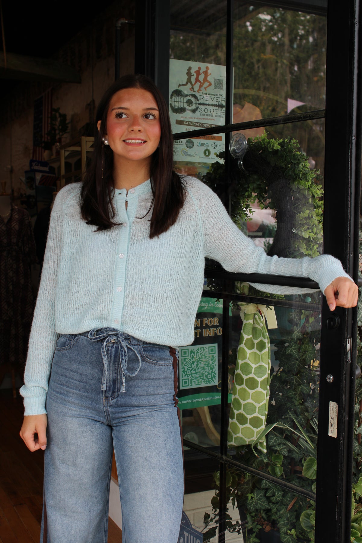 A young woman with dark hair stands in front of a shop window wearing a light blue sweater