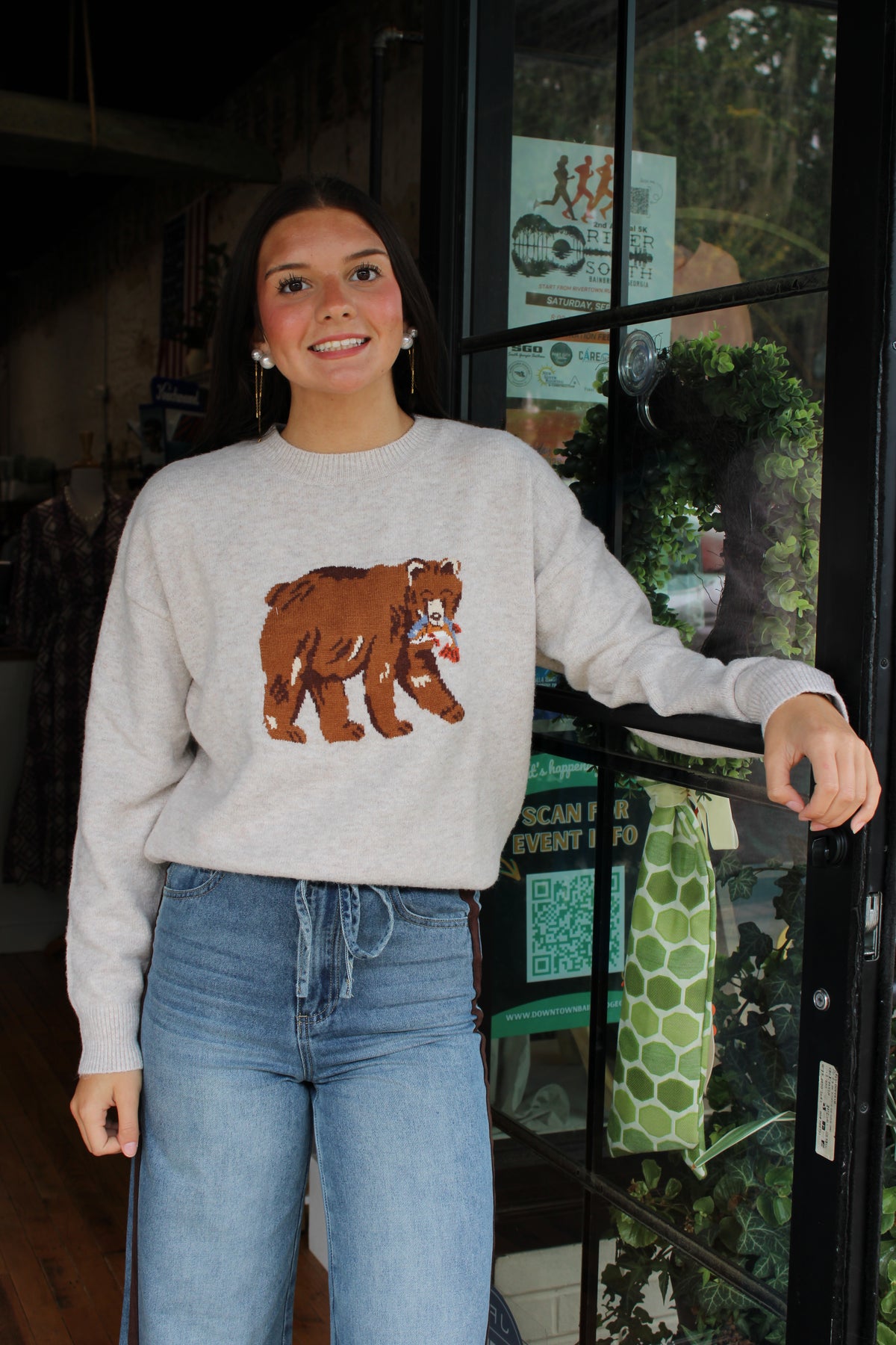 A woman wearing a light gray crewneck sweater with a brown bear graphic on the front, paired with blue jeans, standing in front of a shop window.