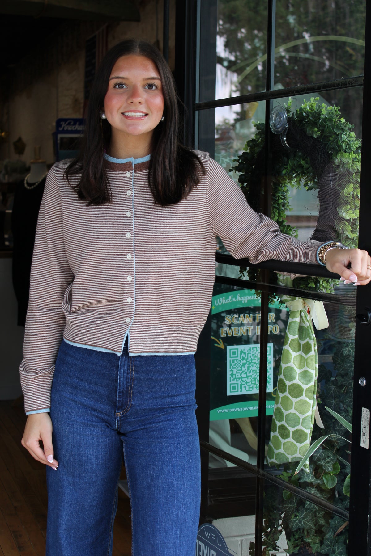 A young woman smiling and posing in front of a storefront window, wearing a beige and white striped button-up cardigan with light blue trim and blue jeans.