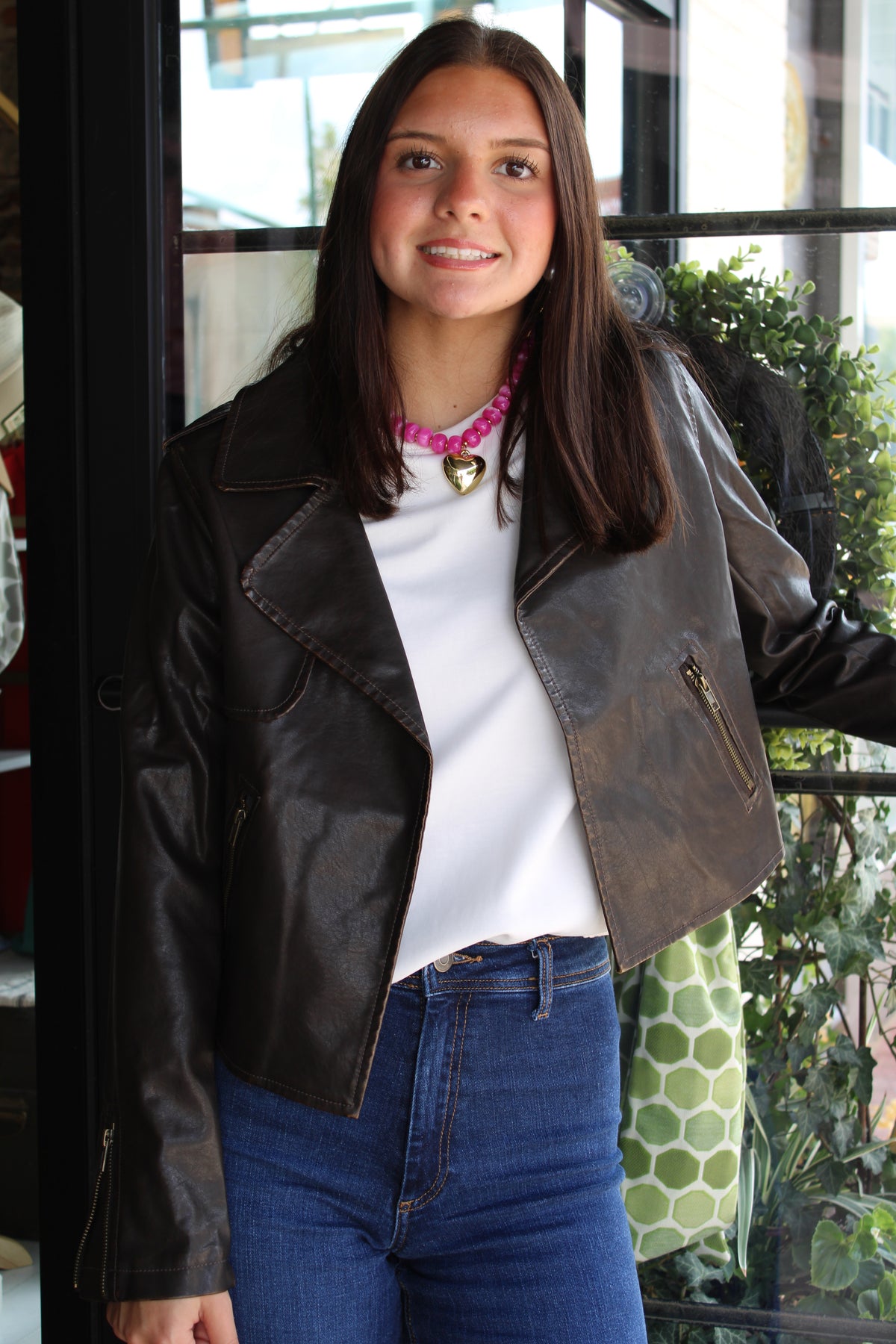 A woman wearing a dark brown distressed leather moto jacket, a white t-shirt, blue jeans, and a pink heart necklace, standing in front of a glass storefront window.