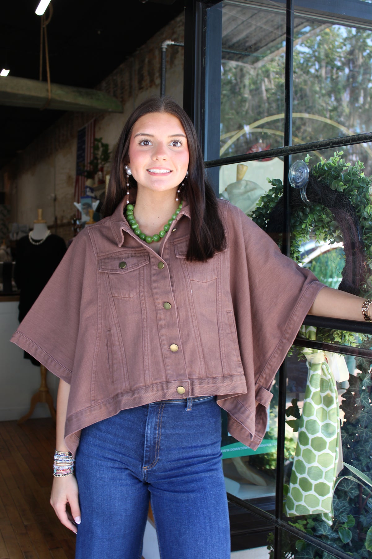 A woman wearing a dusty rose-brown cropped denim poncho-style shirt with blue jeans, standing in front of a shop window.