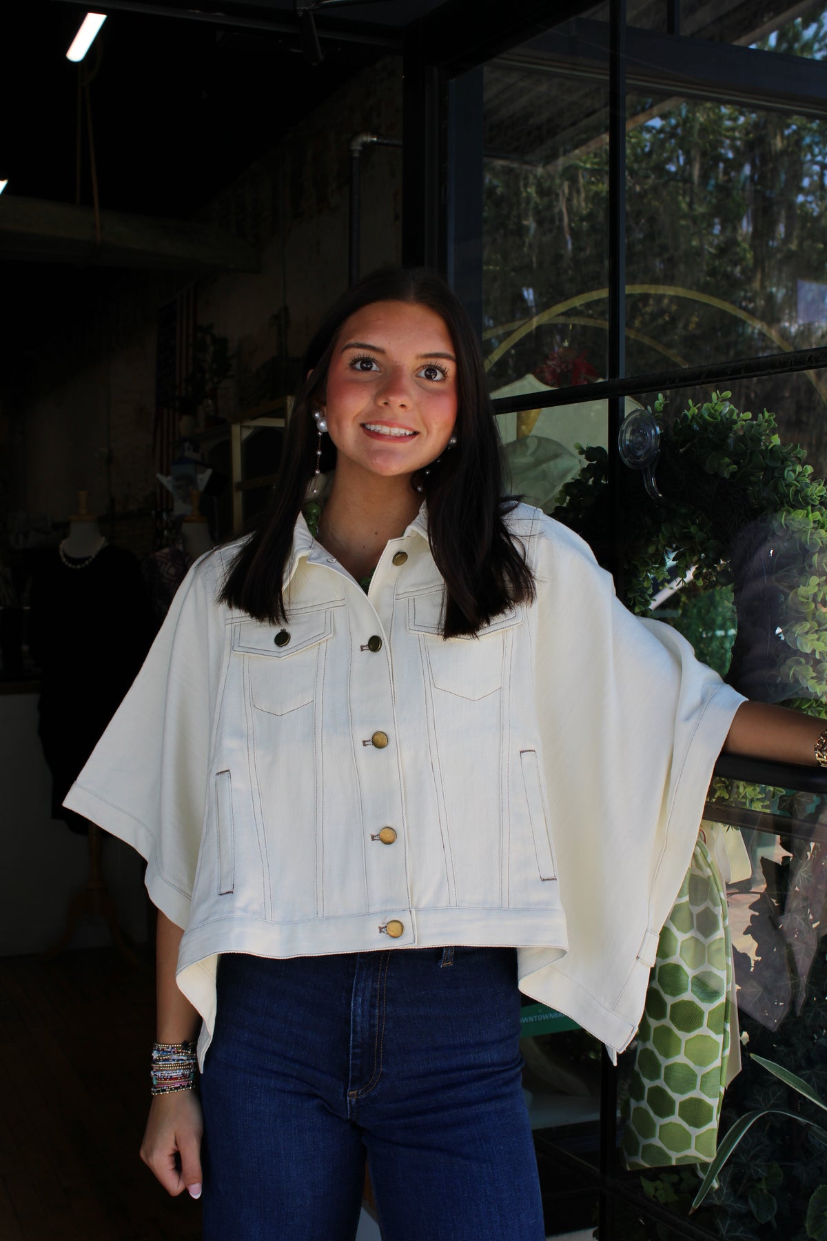 A woman wearing a white denim poncho-style jacket with gold buttons and blue jeans, standing in front of a shop window.