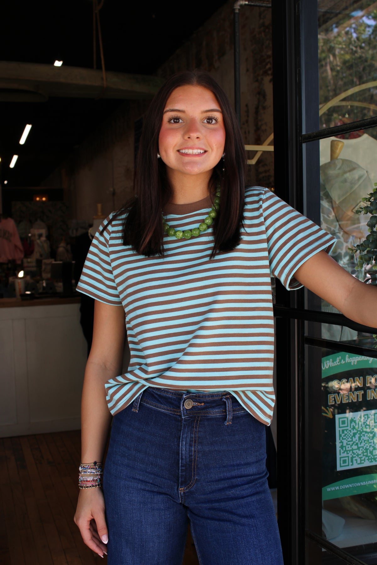 A woman with long dark hair stands indoors wearing a short-sleeve crewneck t-shirt with thin teal and white horizontal stripes, paired with high-waisted blue jeans. She is smiling and posing in front of a glass door of what appears to be a cafe or boutique.