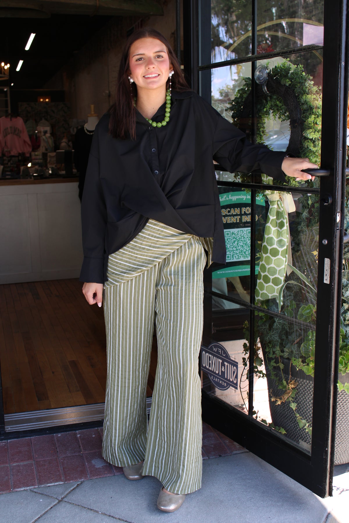 A woman stands in front of a storefront doorway wearing a black long-sleeve top and olive green and white vertically striped wide-leg pants. She is smiling and holding the door frame, with a shop interior and outdoor greenery visible behind her.