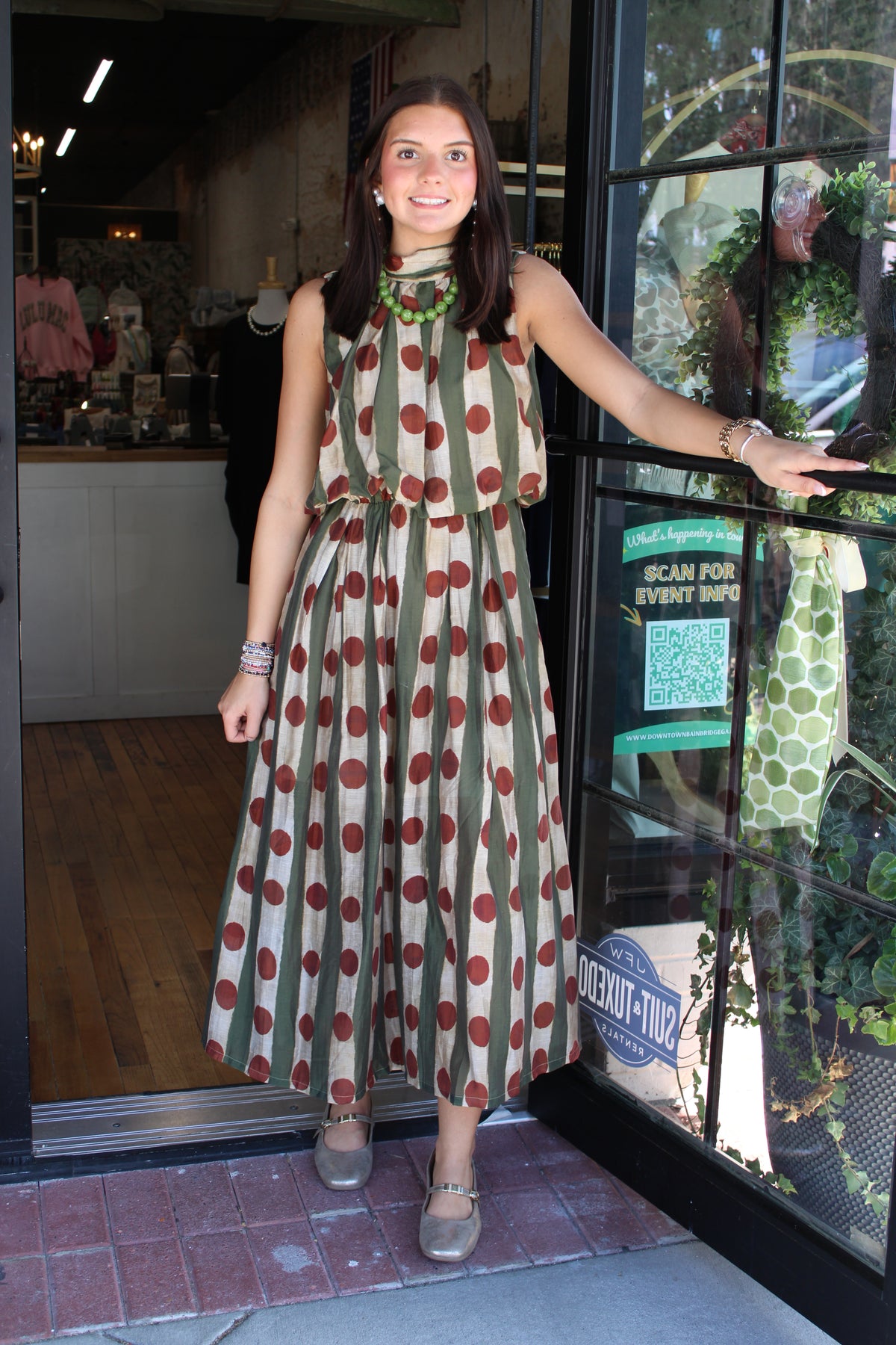 A woman wearing a sleeveless olive green and brown polka dot midi dress stands in the doorway of a boutique, holding the black metal door frame.