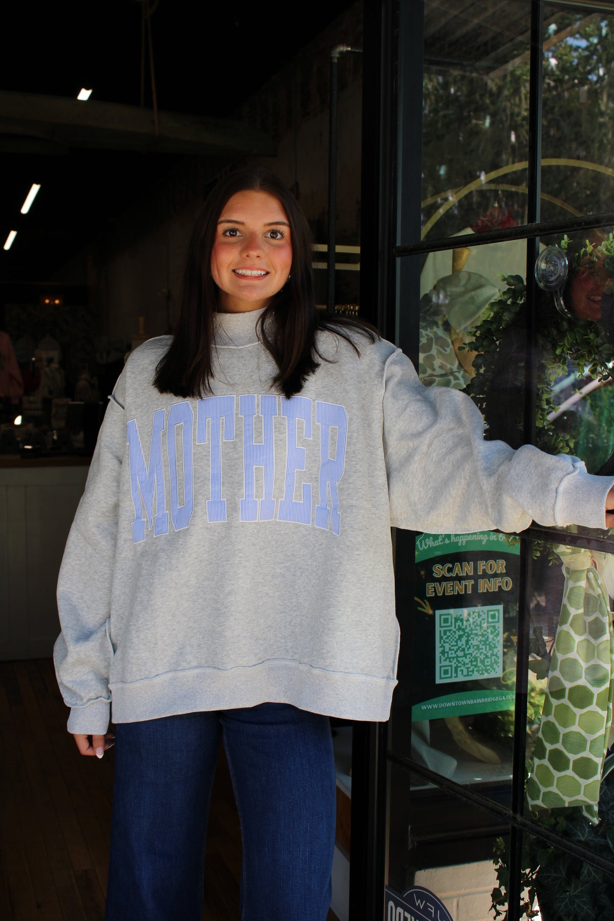 A woman wearing a light gray oversized sweatshirt with 'MOTHER' printed in light blue varsity letters across the chest, standing in front of a glass storefront window.