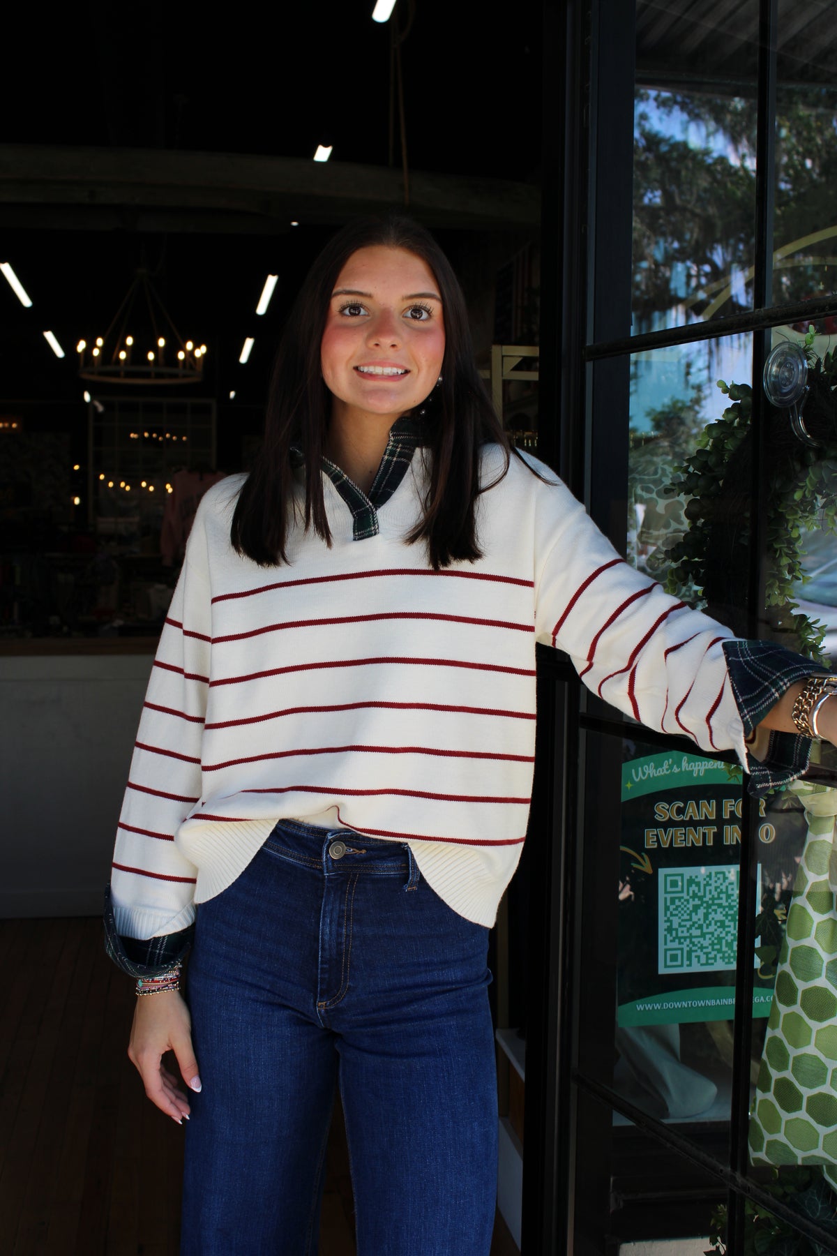 A woman wearing a white knit sweater with thin red horizontal stripes and blue jeans, standing in front of a shop window.