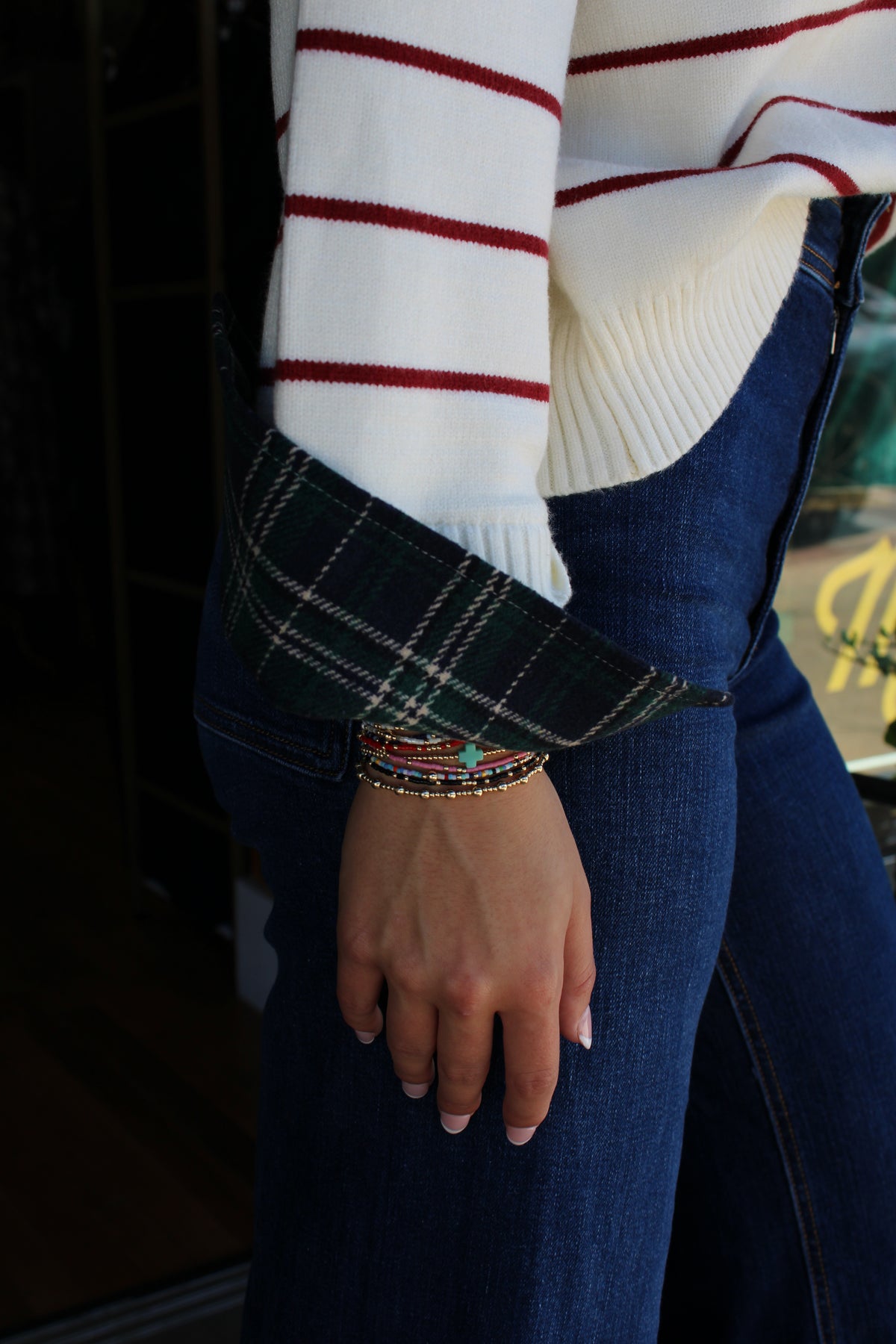 A close-up lifestyle shot of a person wearing a white ribbed sweater with red stripes, a dark green plaid shirt cuff, and blue jeans.
