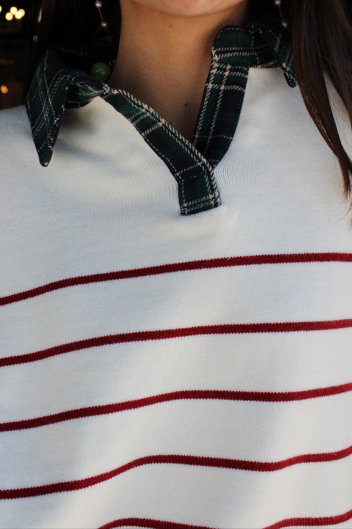 A close-up of a person wearing a white knit sweater with thin red horizontal stripes and a dark green plaid collar detail,