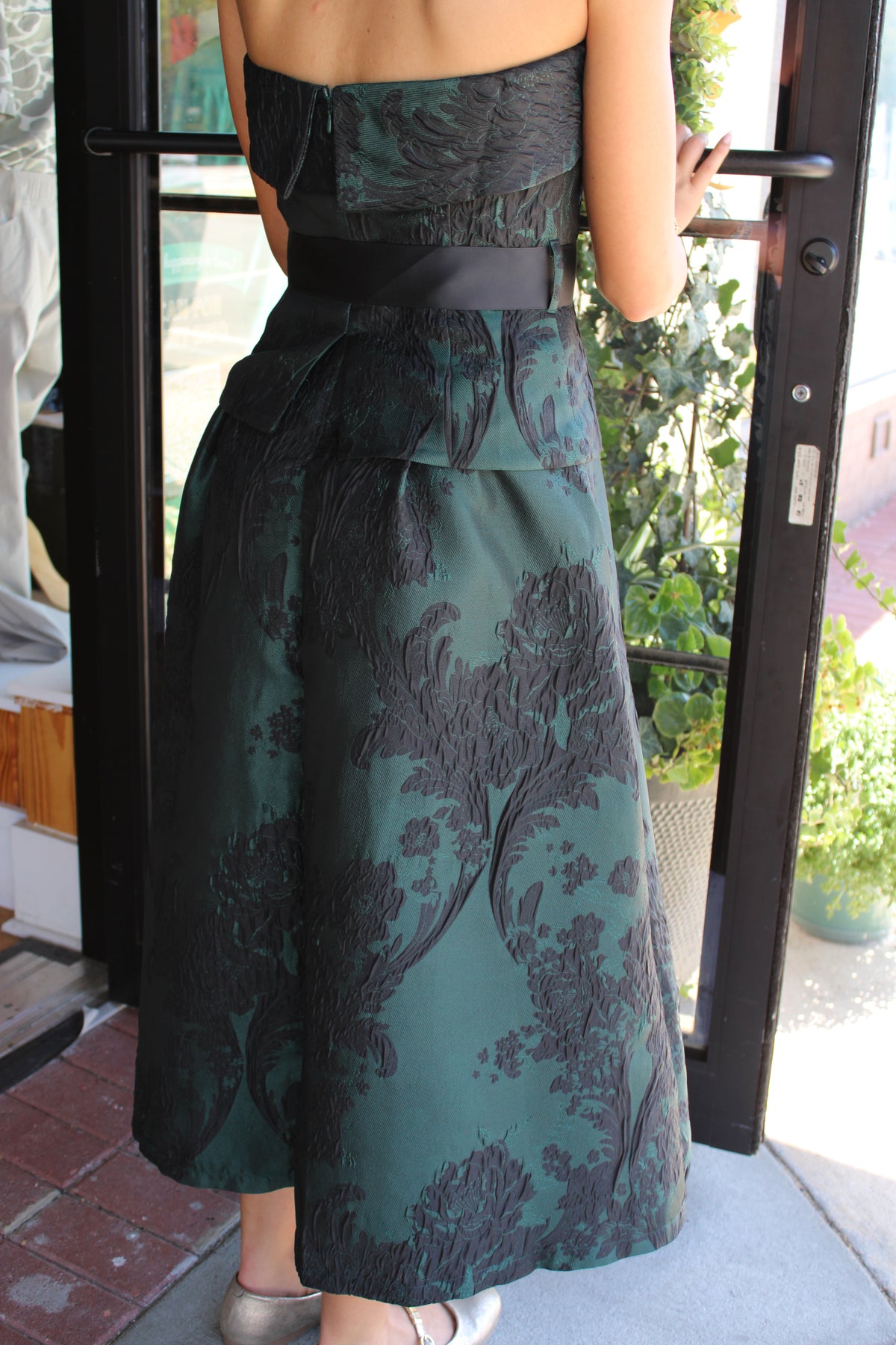 A strapless midi dress with a dark green and black floral jacquard pattern, worn by a model standing at a glass door.