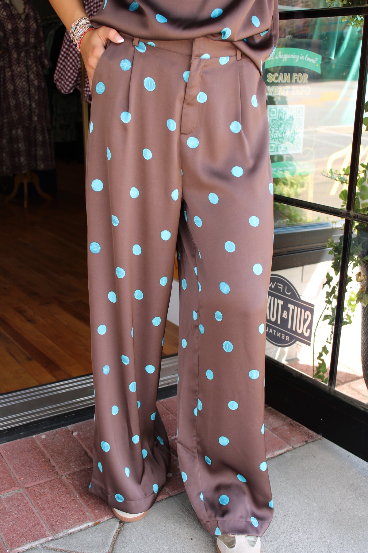 A person wearing wide-leg brown pants with a light blue polka dot pattern, standing on a sidewalk in front of a storefront window.