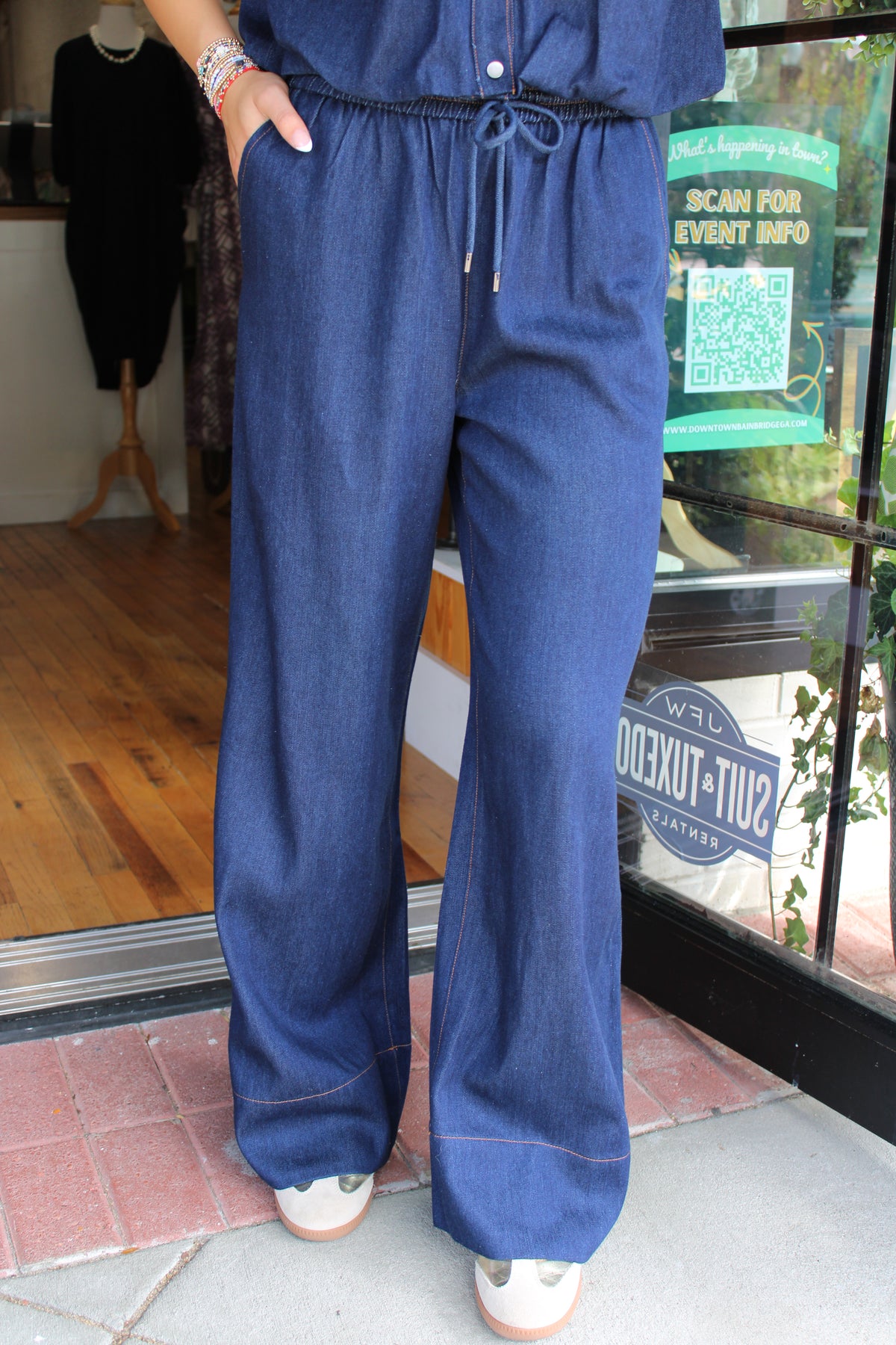 A person wearing a dark blue denim pants with wide legs and a drawstring waist, standing on a sidewalk in front of a storefront window.