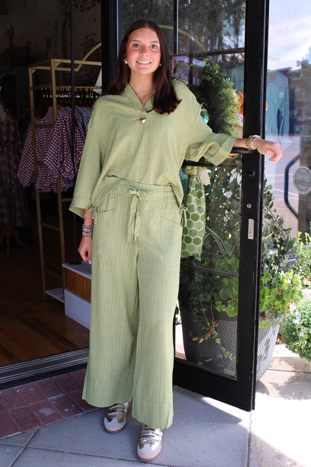 A woman stands in a storefront doorway wearing a light sage green pinstriped matching set consisting of a long-sleeve button-front top and wide-leg drawstring pants. She is smiling and posing with her hand resting on the doorframe.