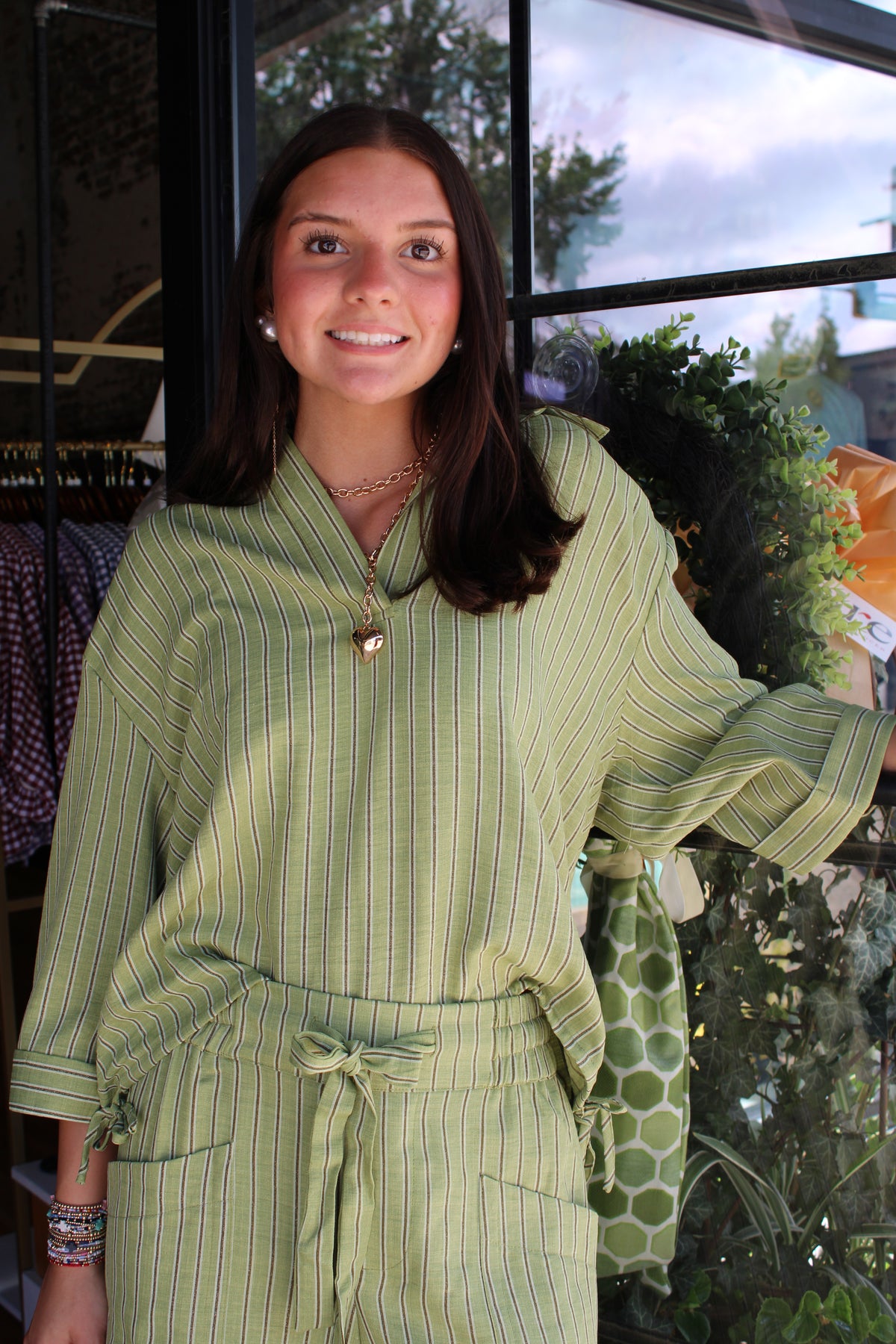 A woman wearing a light green and white vertically striped long-sleeve top with a tie waist, standing in front of a window.