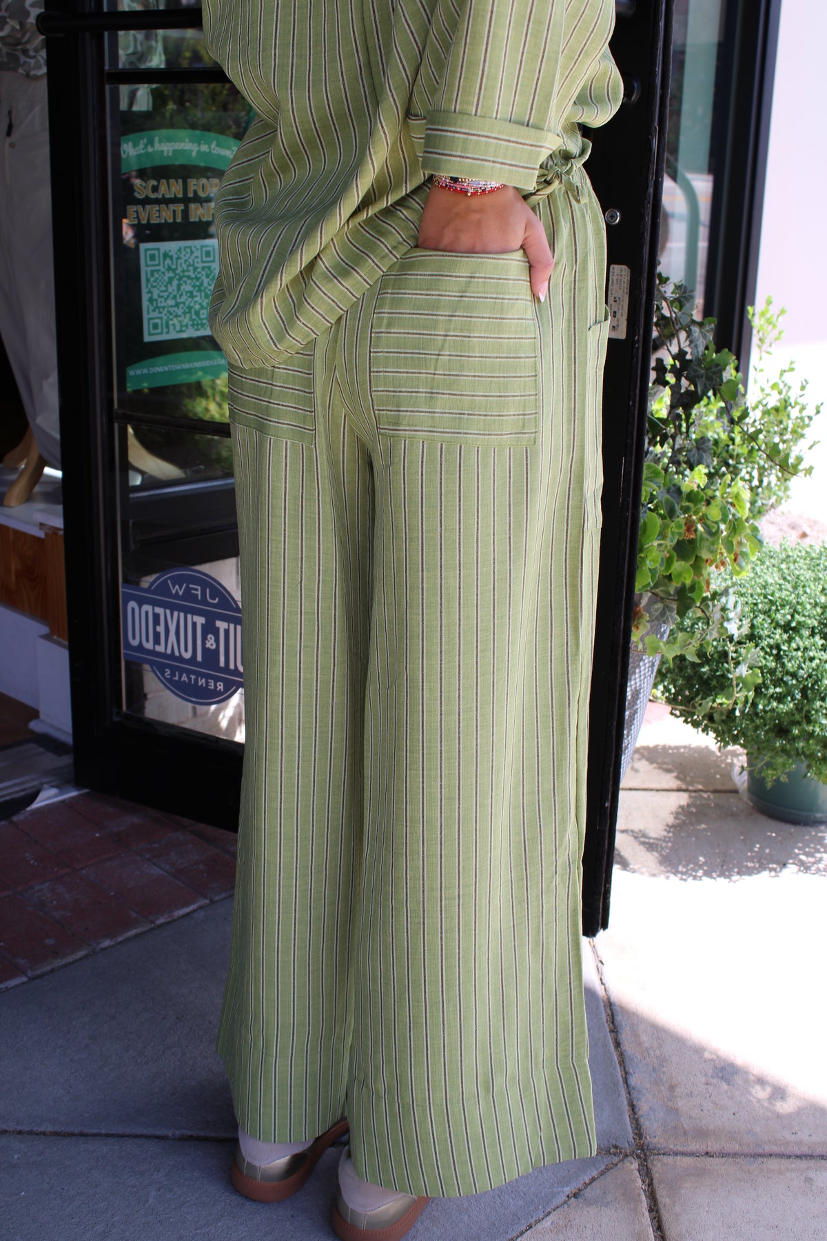A person wearing a light green and white vertically striped matching set consisting of a long-sleeve top and wide-leg pants, standing on a sidewalk in front of a storefront.