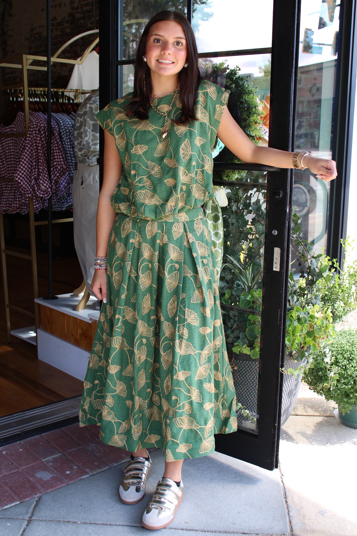 A woman wearing a green sleeveless top and matching skirt with a gold leaf pattern, standing in front of a glass storefront door.