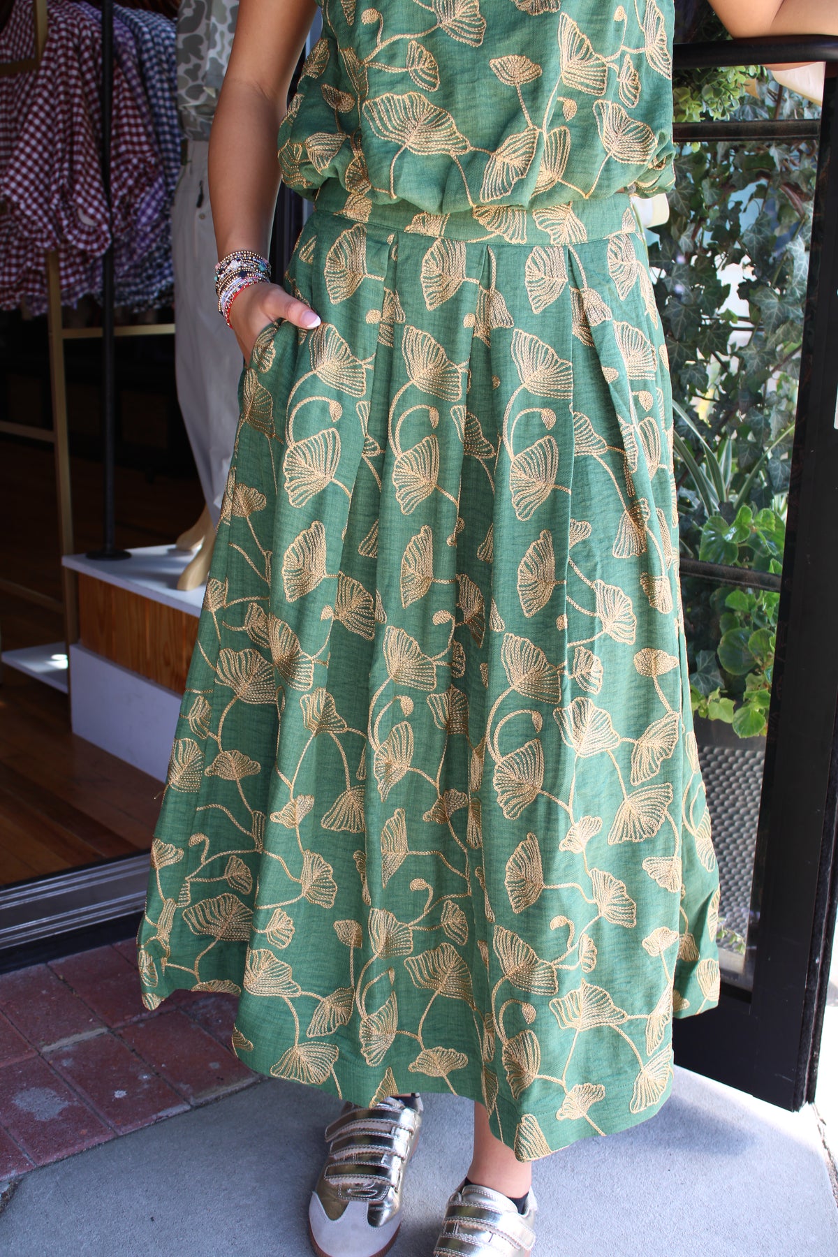 A person wearing a green skirt with a beige leaf pattern, standing in front of a glass door.