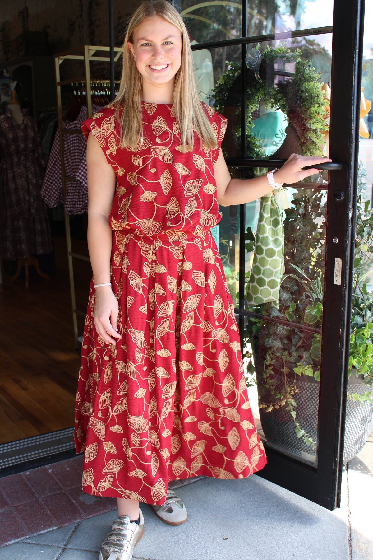 A woman stands in a storefront doorway wearing a sleeveless red top and matching skirt with a gold leaf pattern. She is holding the door open with one hand, with a clothing rack and shop interior visible behind her.