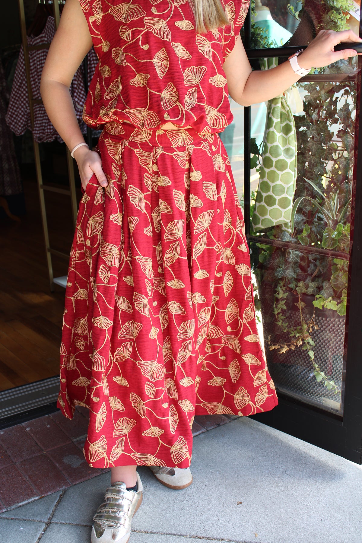A person wearing a sleeveless red skirt with a gold leaf pattern, standing in a doorway. The dress has a gathered waist and a flared skirt, paired with white sneakers.