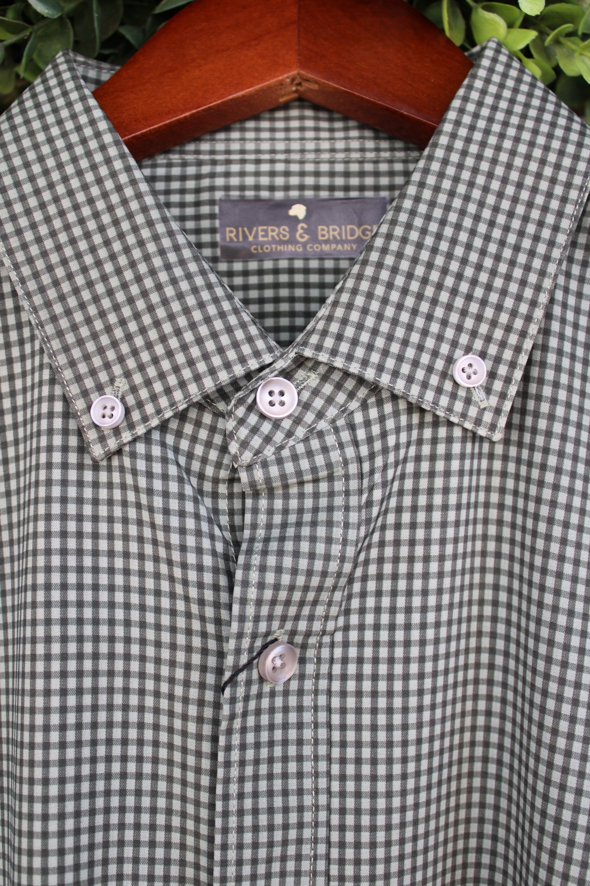 A close-up of a Rivers & Bridge button-down shirt in green and white, hanging on a wooden hanger with green foliage in the background.