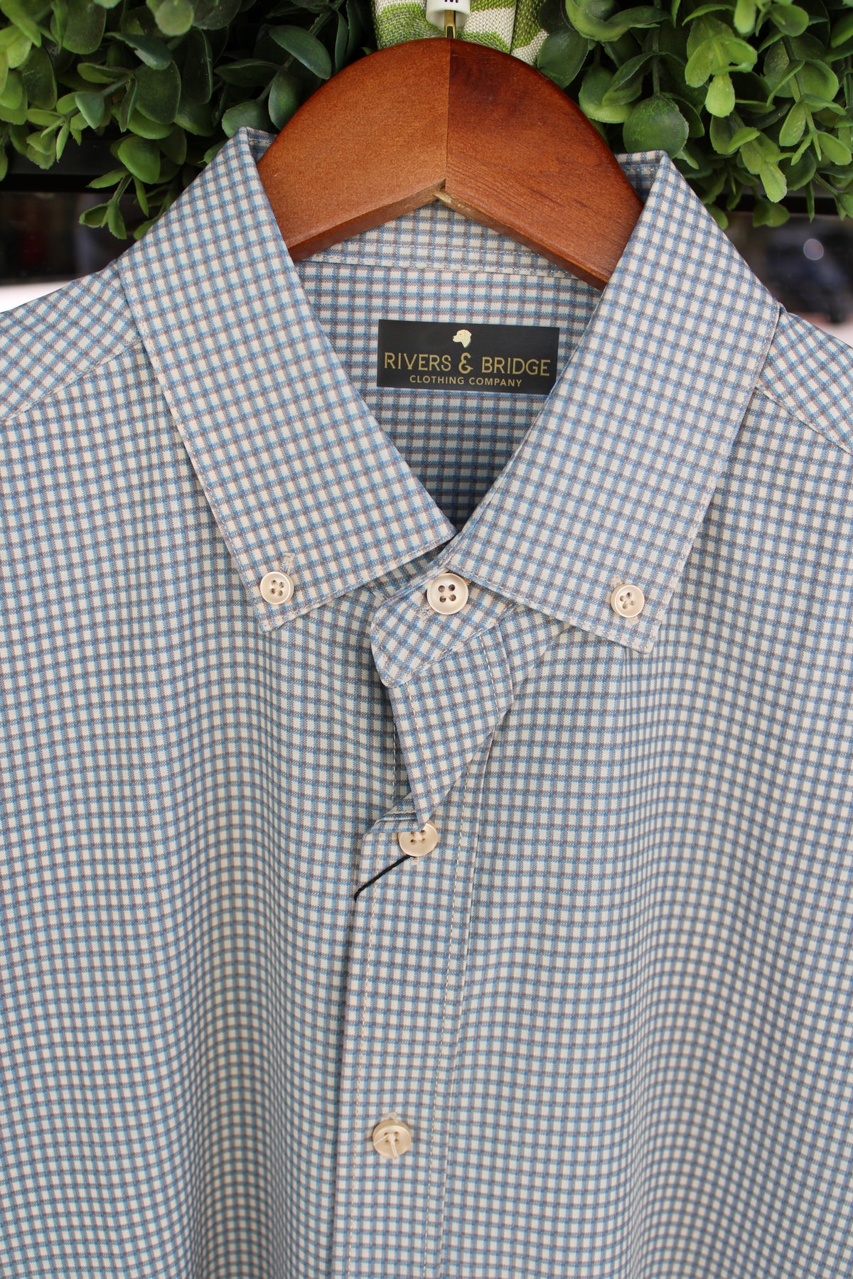 A light blue and white button-down shirt by Rivers & Bridge, hanging on a wooden hanger against a background of green foliage.
