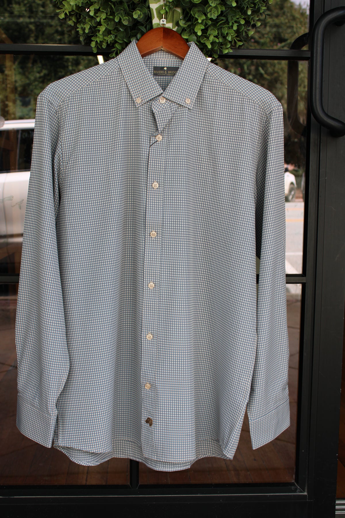 A long-sleeve button-down shirt with a small blue and white checkered pattern, hanging on a wooden hanger in front of a window.