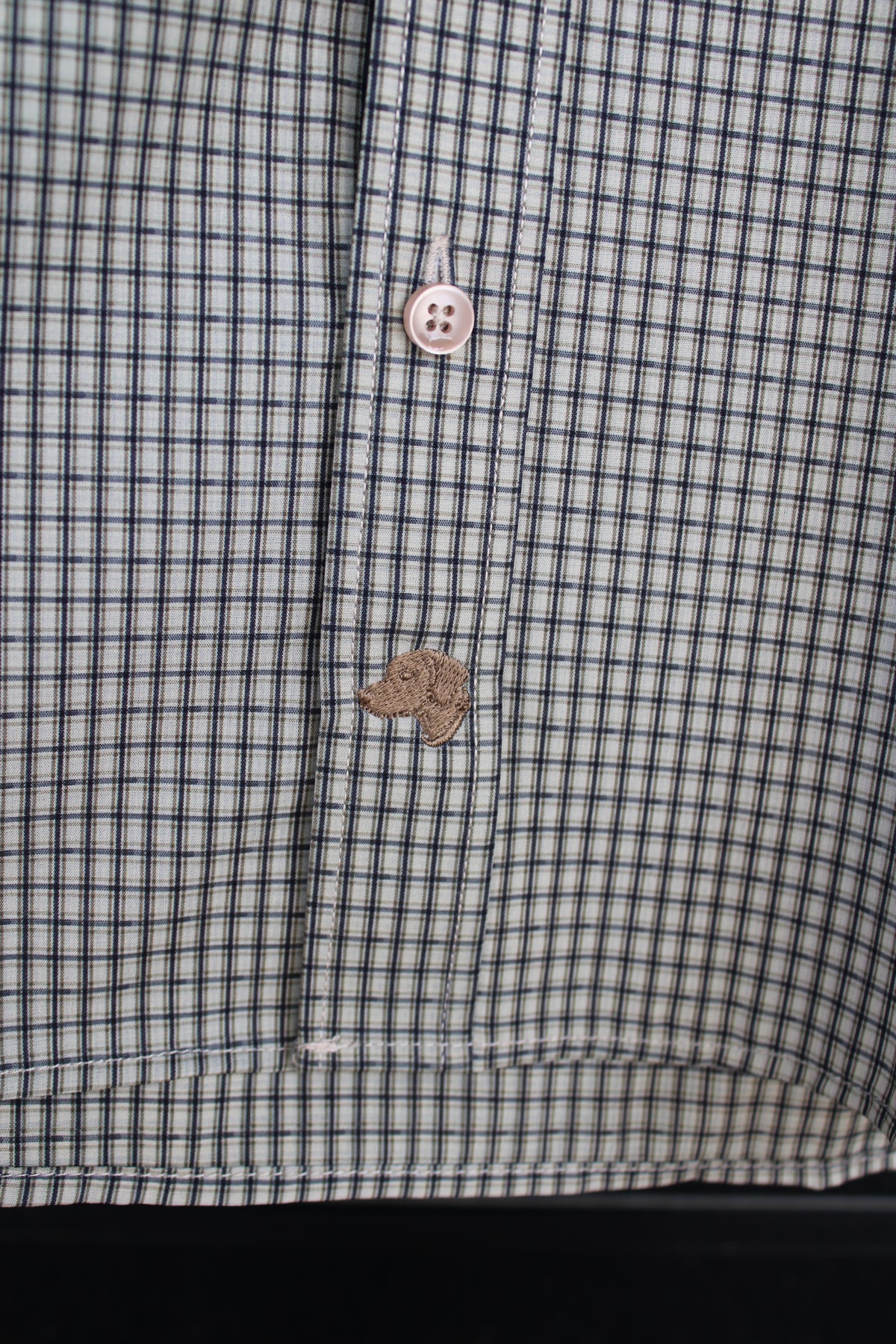 Close-up of a button-down shirt with a fine blue and white gingham pattern, featuring a light pink button and a brown embroidered paw print on the pocket flap.