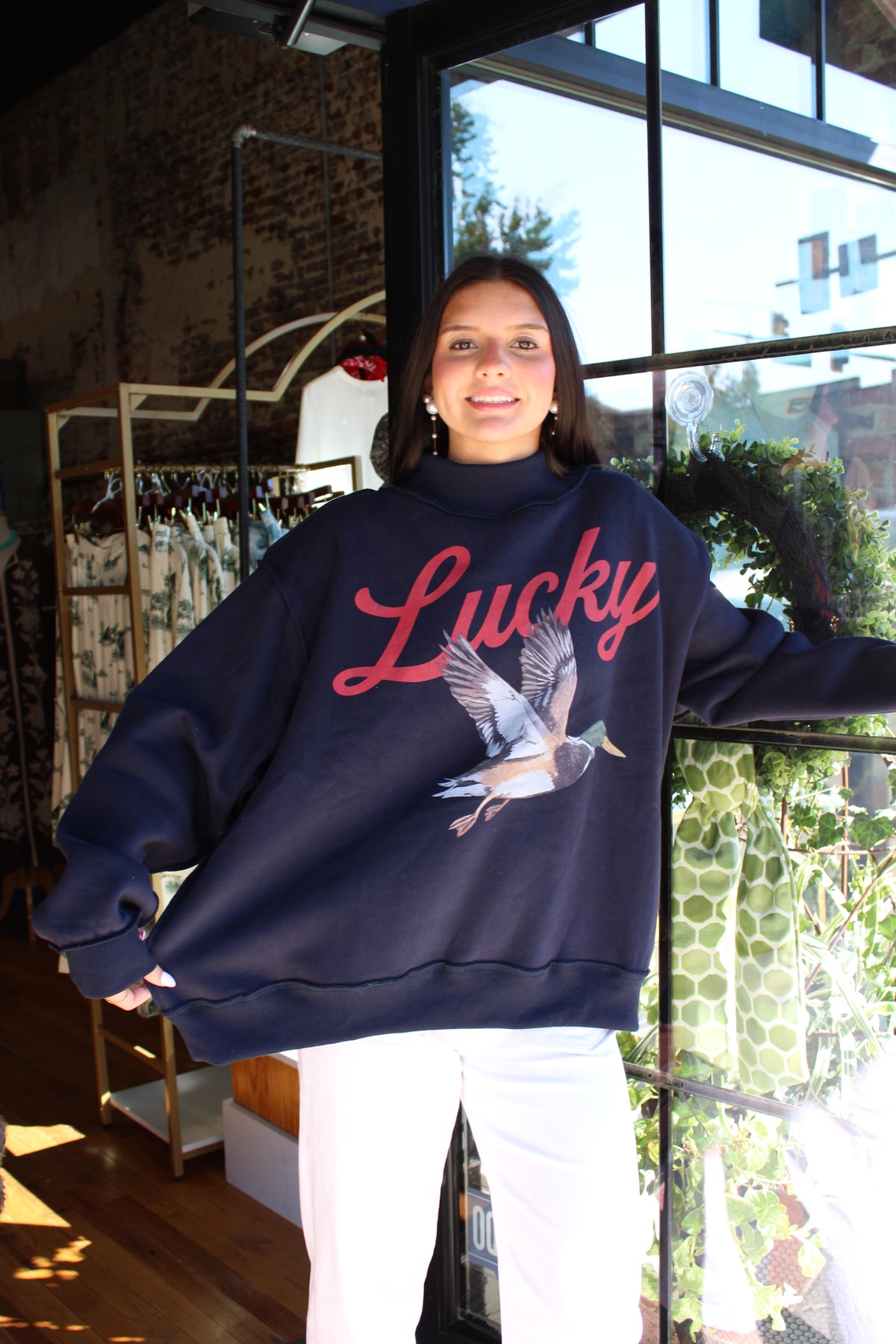 A woman wearing an oversized navy blue sweatshirt with a red 'Lucky' script and a mallard duck graphic, paired with white pants, standing in a shop in front of a large window.