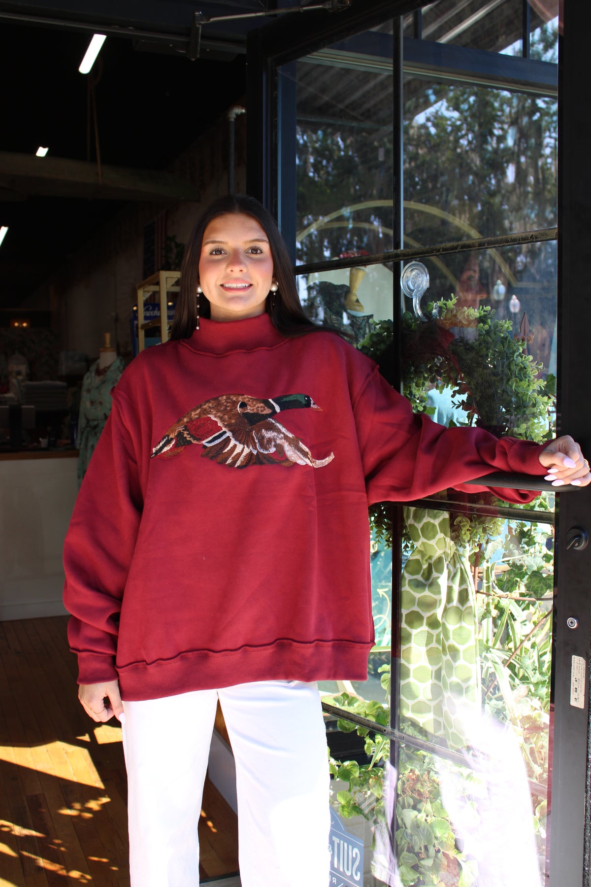 A woman wearing a burgundy turtleneck sweatshirt with a detailed embroidered bird design on the chest, paired with white pants, standing in front of a glass storefront window.
