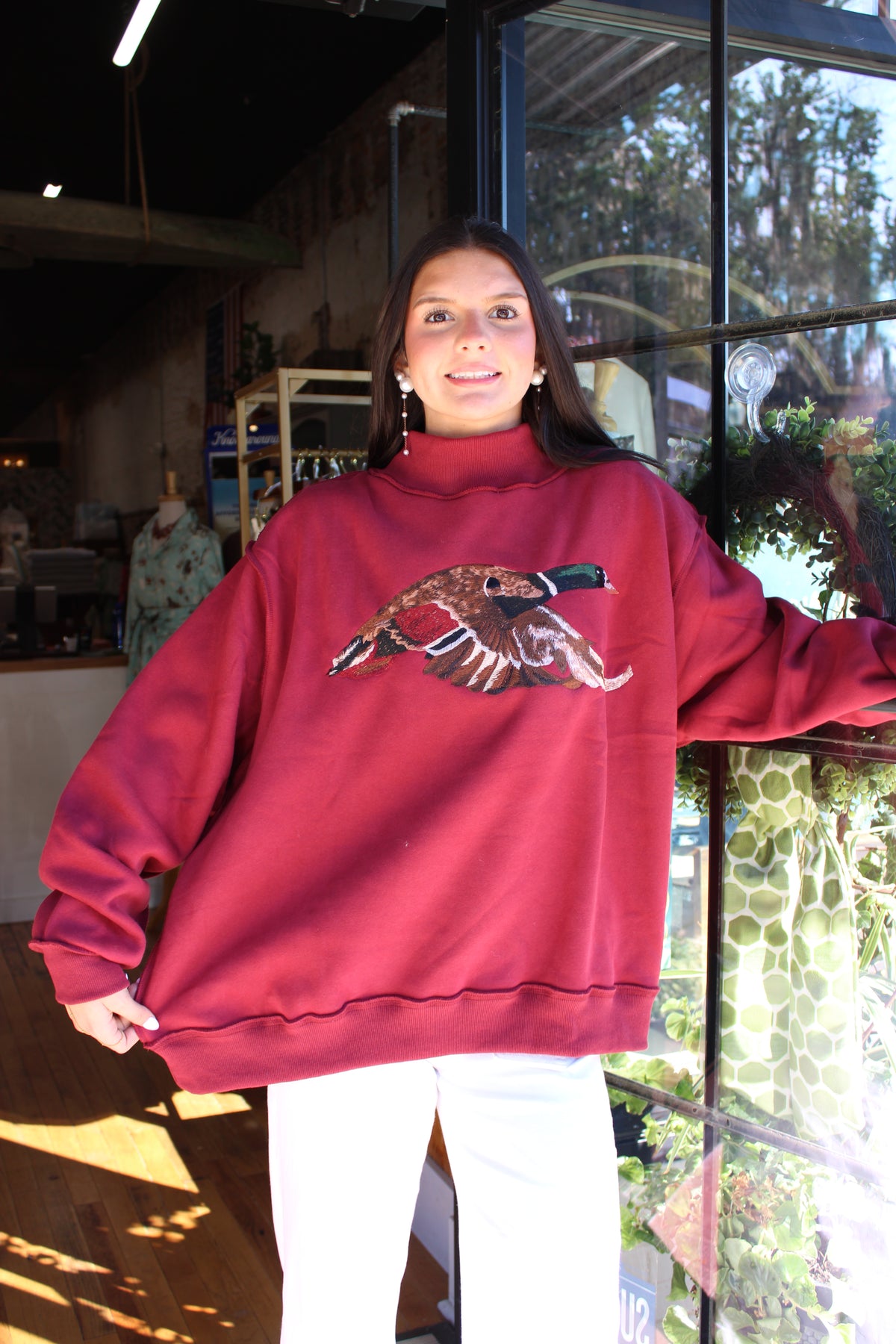 A woman wearing an oversized burgundy sweatshirt with a detailed duck graphic on the chest, paired with white pants, standing in front of a shop window.