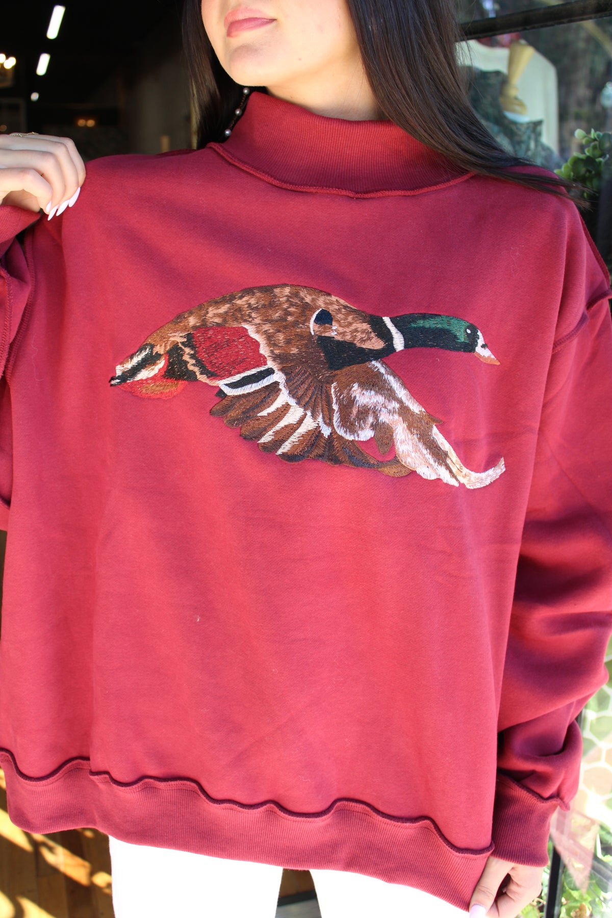 A woman wearing a dark red mock-neck sweatshirt featuring a large, detailed embroidered flying duck on the chest.