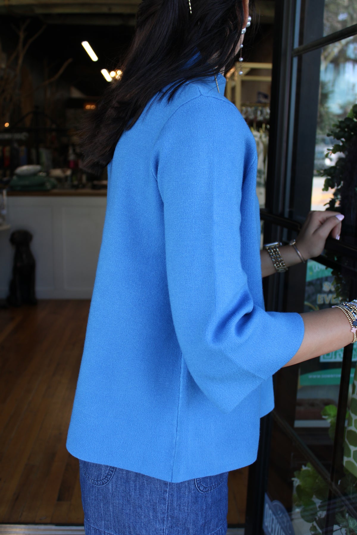 A woman wearing a bright blue three-quarter sleeve sweater and blue jeans, standing in a shop next to a glass door.