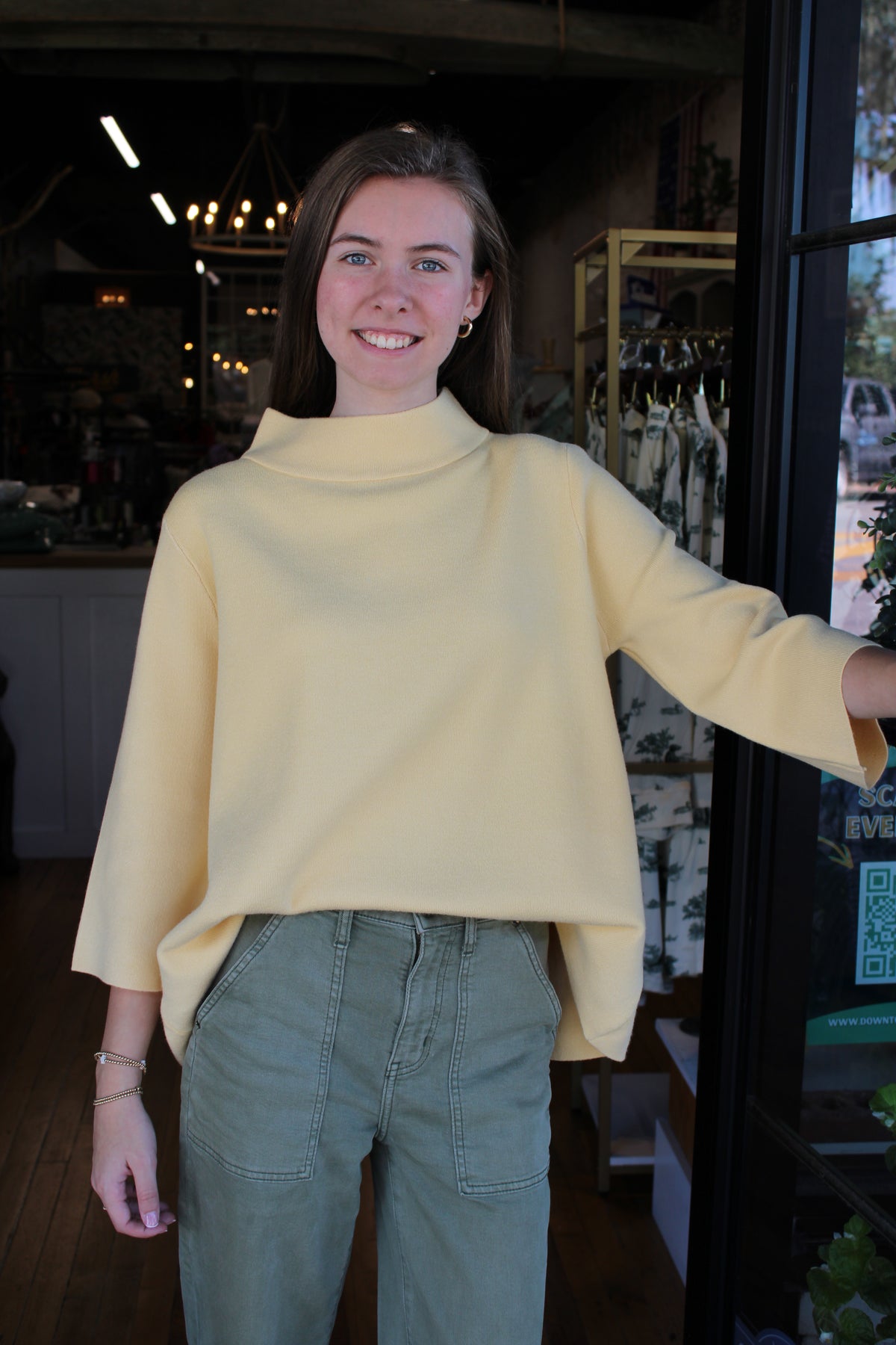 A woman wearing a pale yellow mock-neck long-sleeve top and olive green pants, standing in a shop doorway.