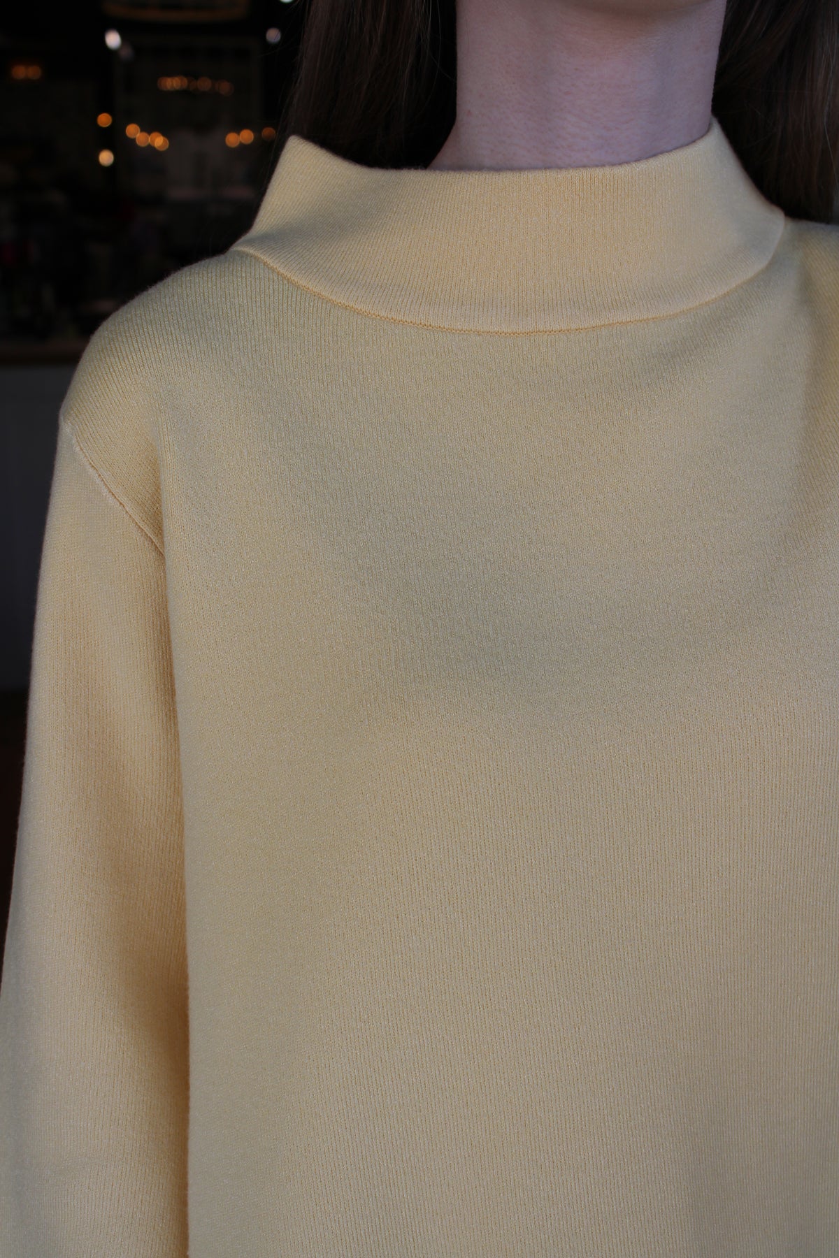 A close-up of a person wearing a pale yellow mock-neck knit top, showing the collar and upper chest against a dark, softly blurred background.