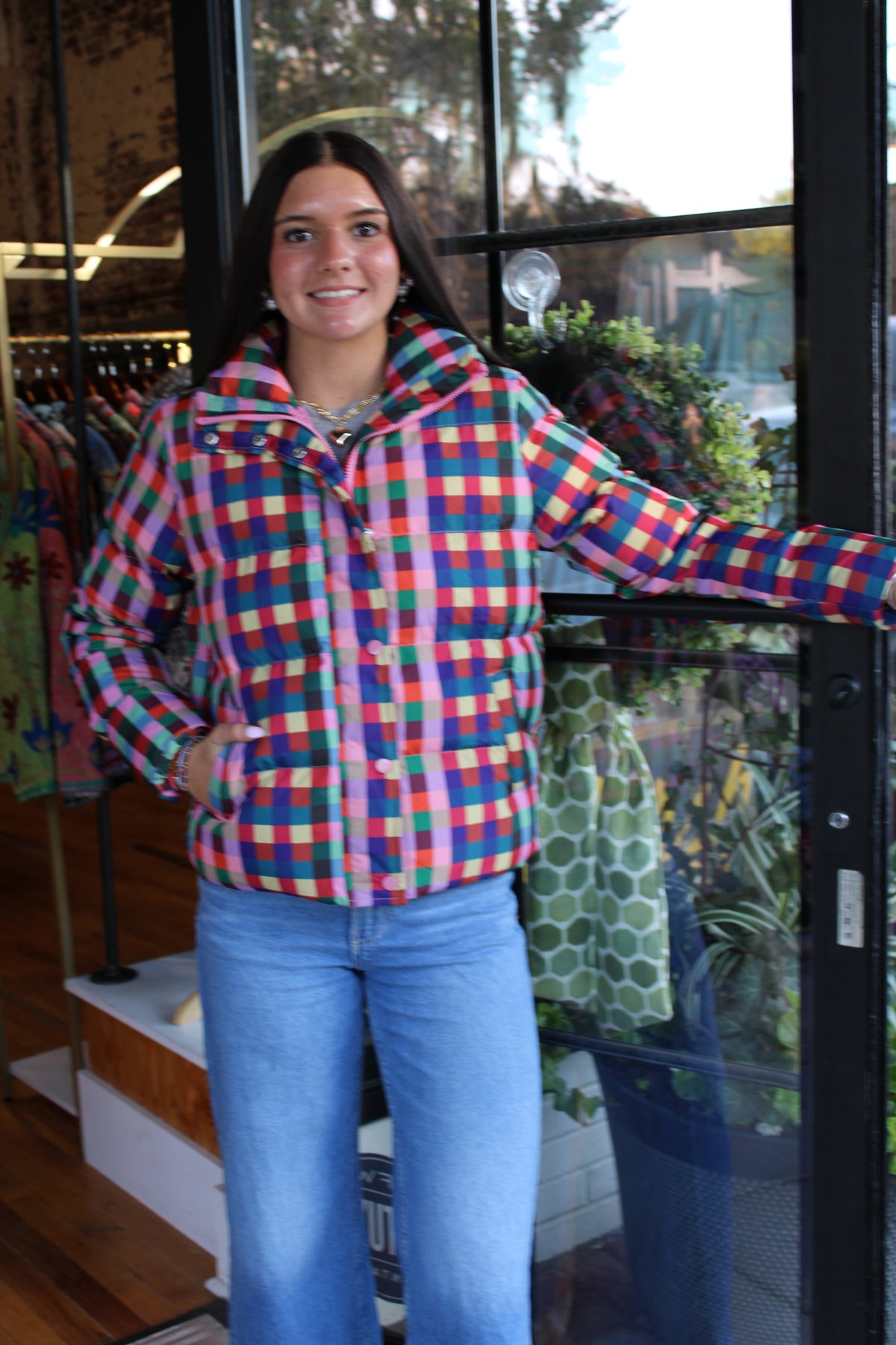 A woman wearing a colorful plaid zip-up jacket and blue jeans stands in a boutique doorway, smiling at the camera.