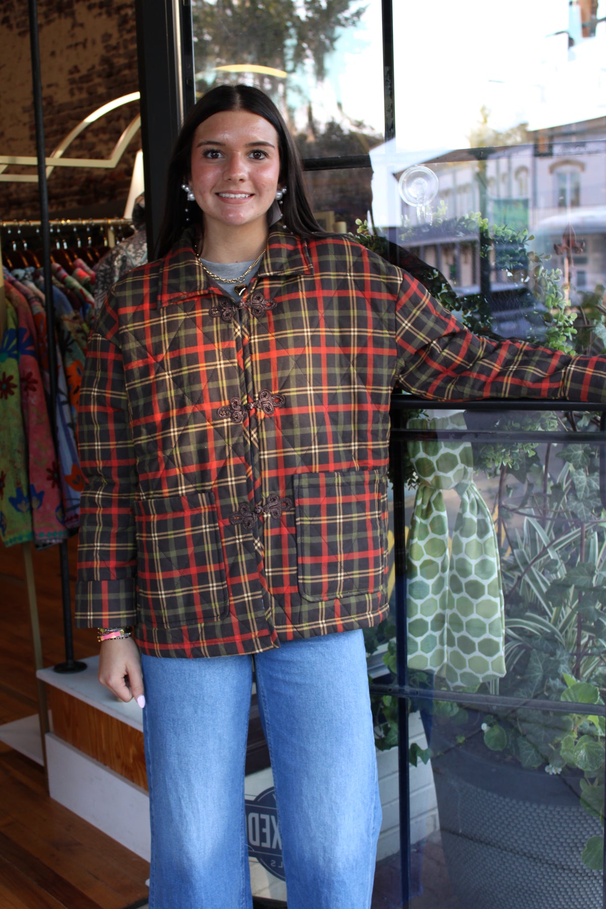 A woman wearing a vintage red, yellow, and black plaid button-up jacket with toggle closures, paired with blue jeans, standing in a clothing boutique.